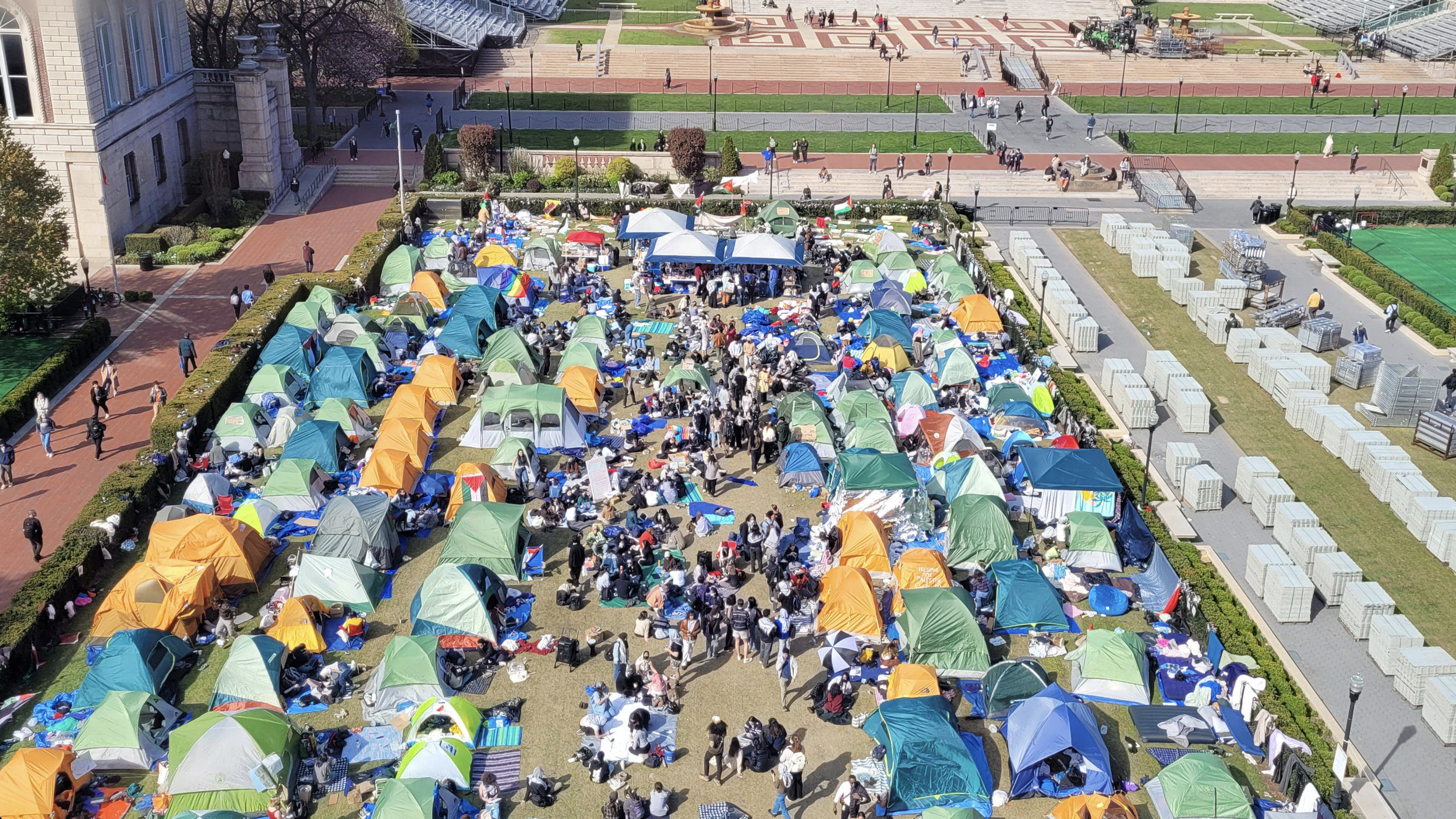 A large outdoor gathering with numerous tents and people, set in a city park or plaza, with pathways, greenery, and a multi-level ledge in the background.