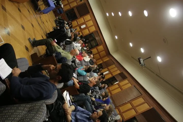 A large group of people seated in rows of chairs inside a conference room or lecture hall, listening to a presentation or speaker, with some taking notes.