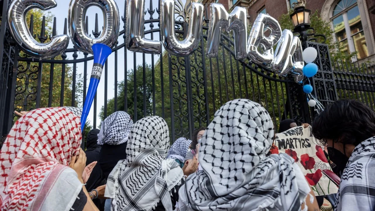 A group of people wearing Palestinian scarves gather outside a gate decorated with silver balloons that spell 'JOUJOUR,' with some holding signs with red poppies and the words 'Honor Martyrs.'