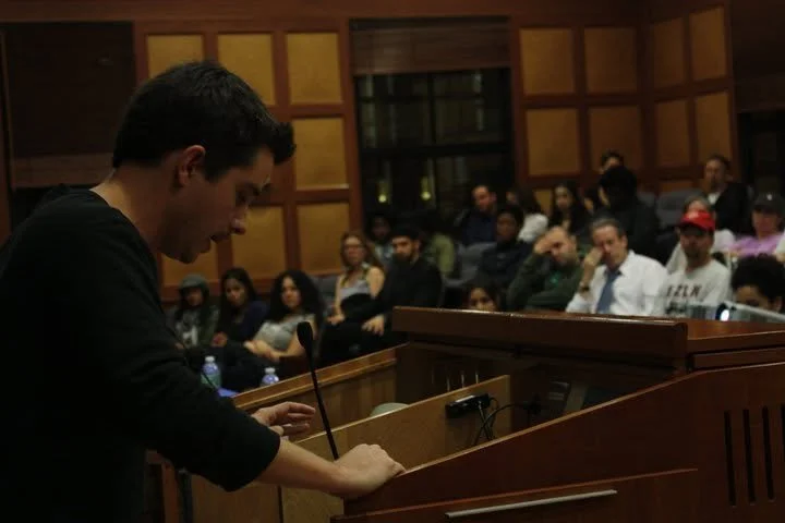 A young man speaking at a podium in a courtroom or meeting room with an audience seated behind him.