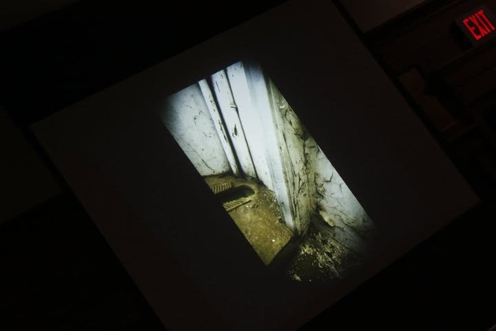 Dark, dirty corner of an abandoned building with peeling paint, grime, and debris, illuminated by a faint light from a doorway.