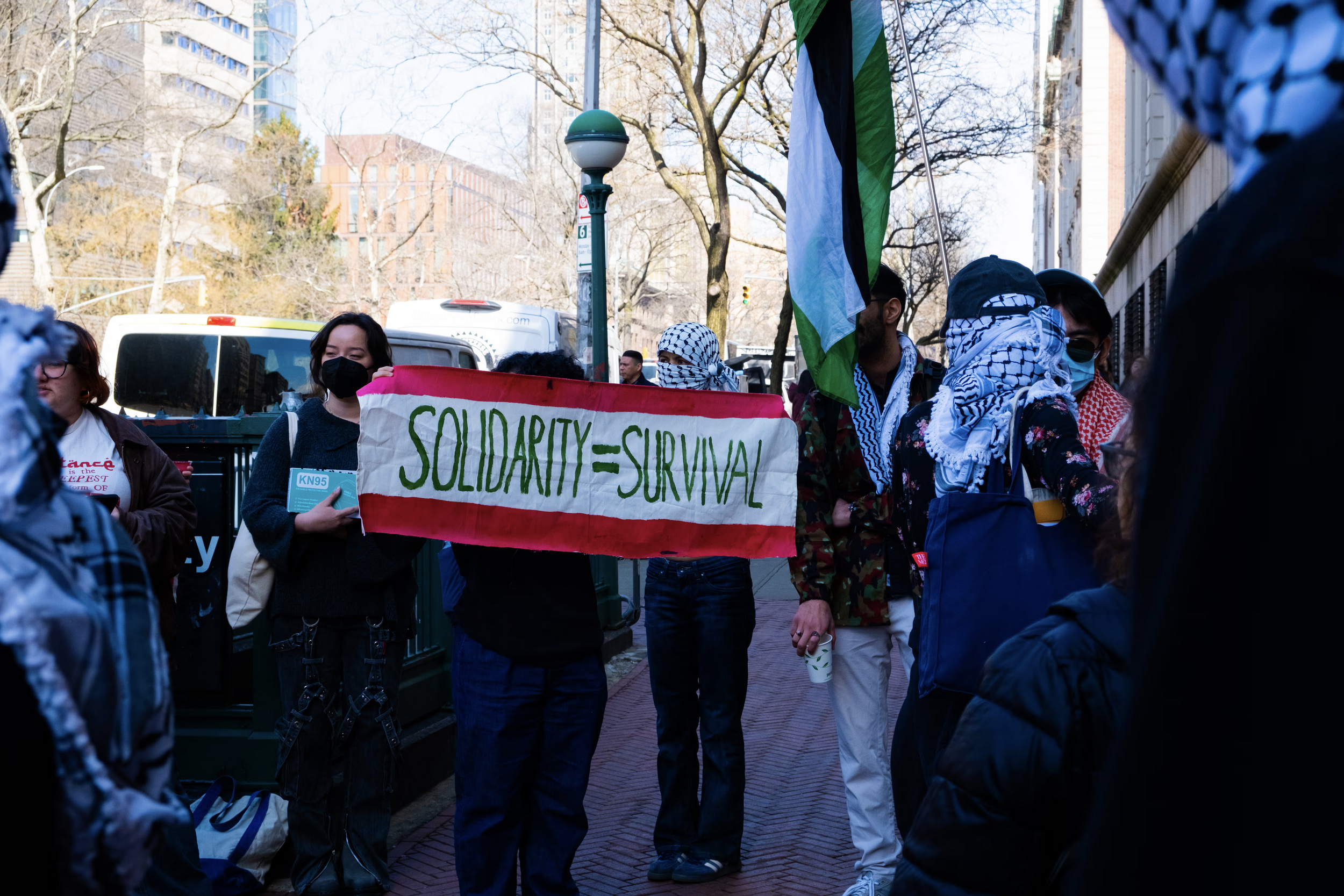 Group of protesters wearing masks and holding a sign that reads 'SOLIDARITY = SURVIVAL' on a city street with buildings and trees in the background.