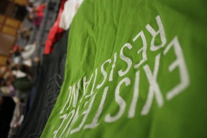 Green fabric with white letters spelling 'CXSISC' and more text, blurred background of people and objects.
