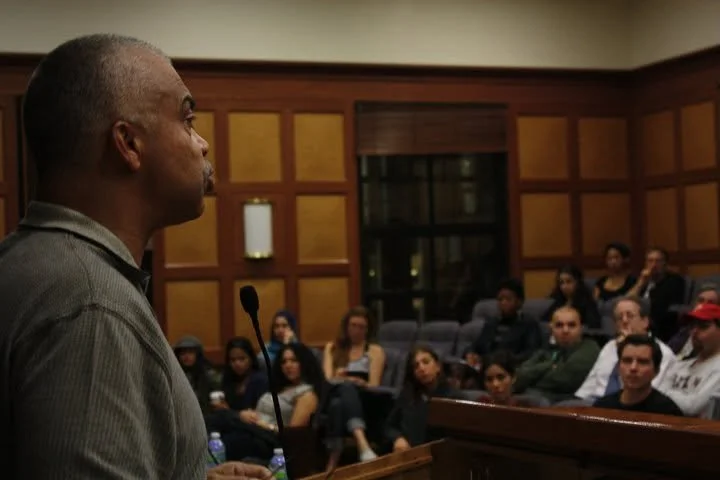 Man speaking at a podium in a courtroom with an audience seated behind him.