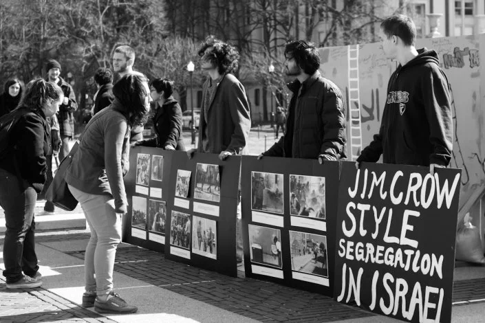 People at an outdoor protest or demonstration, displaying signs and photographs, with a large sign reading 'JIM CROW STYLE SEGREGATION IN ISRAEL'.