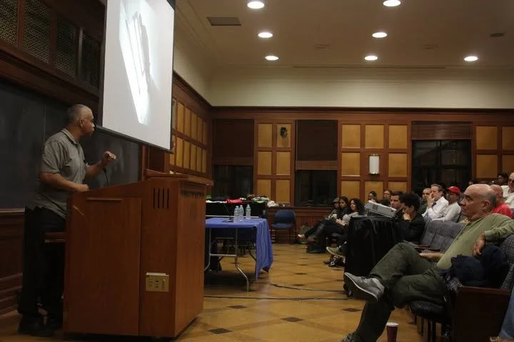 A man giving a presentation in a wood-paneled conference room filled with seated audience members.