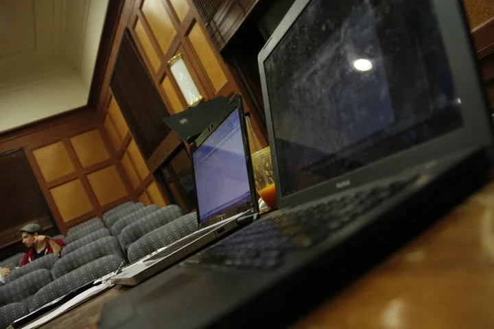 Multiple laptops on a wooden table in a room with wood-paneled walls, a row of gray upholstered chairs, and a person sitting in the background.