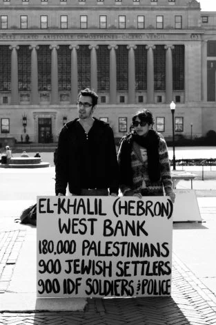 Two people holding a protest sign on a city street. The sign reads: 'EL-KHALIL (HEBRON) WEST BANK, 180,000 PALESTINIANS, 900 JEWISH SETTLERS, 900 IDF SOLDIERS? POLICE.' In the background, there is a large historic building with columns.