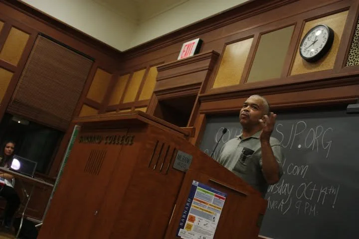 A man standing at a podium giving a speech in a wood-paneled room, with a chalkboard behind him, and a woman sitting at a table with a laptop in the background.