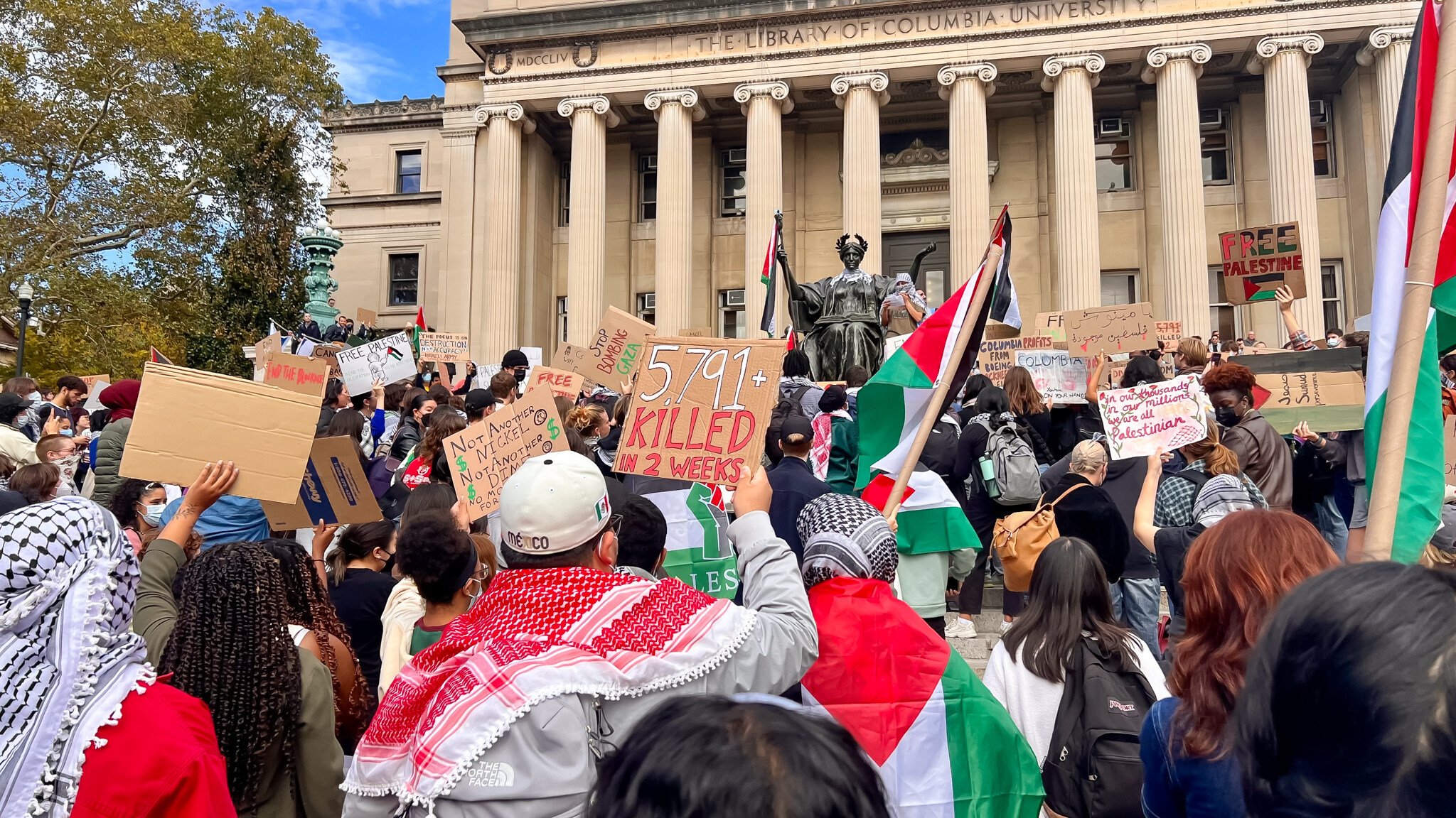 A large crowd of protesters gathers outside a building with tall columns, holding various signs and Palestinian flags during a demonstration. The signs include messages like '5,791+ Killed in 2 Weeks,' 'Free Palestine,' and other political statements. Many participants are wearing masks and traditional headscarves.