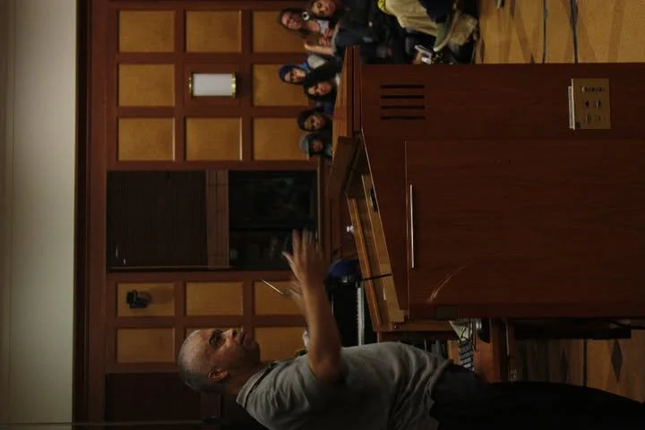 A man giving a presentation or speech at a podium in a wood-paneled room, with a group of people seated in the background.