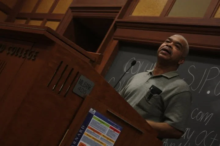 A man wearing a gray polo shirt standing behind a wooden podium in front of a chalkboard in a classroom or lecture hall. He appears to be speaking or giving a presentation.