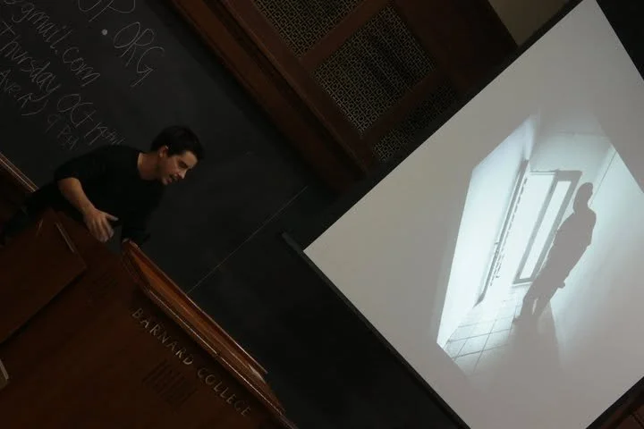 A woman is standing at a podium in front of a blackboard at a college classroom, smiling and holding a phone. There is a projection of a black and white photo of a person standing near a door in a hallway on the screen behind her.