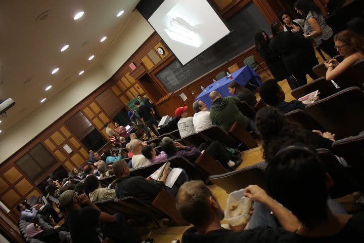 A classroom or lecture hall filled with people attending a presentation. Some are seated, reading or listening, while others stand near the back and side. There is a chalkboard at the front, a projector screen displaying an image, and tables with blu