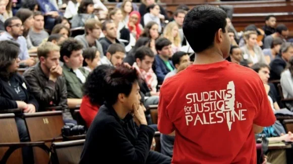 A person wearing a red shirt with the text "Students for Justice in Palestine" standing in front of a large classroom or lecture hall filled with students.