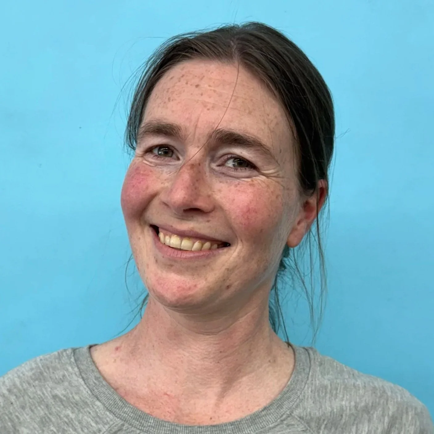 A smiling woman with brown hair and freckles, wearing a grey shirt, standing against a light blue background.