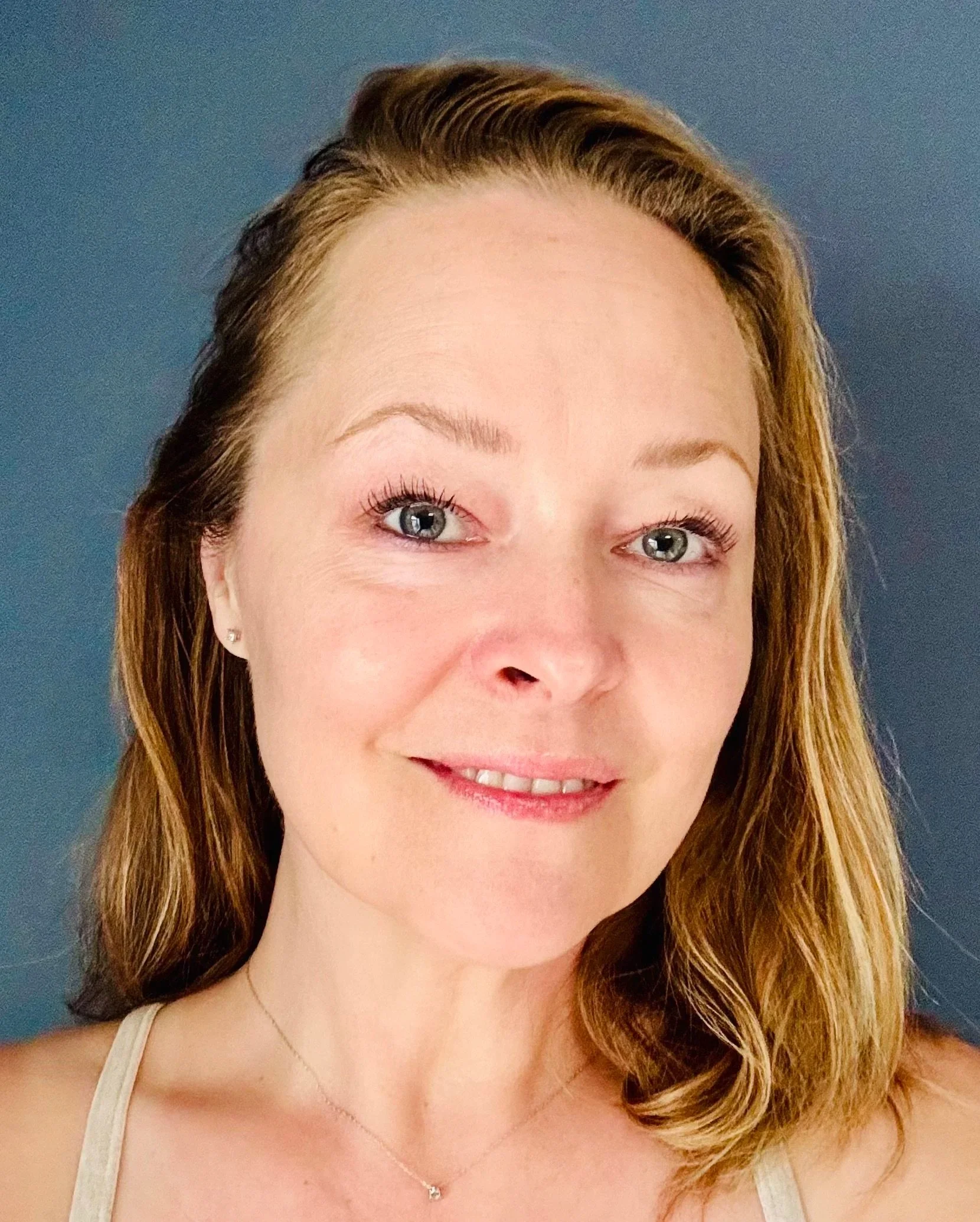 Close-up of a woman with long light brown hair, blue eyes, and wearing a light-colored tank top and a small necklace, smiling in front of a blue background.