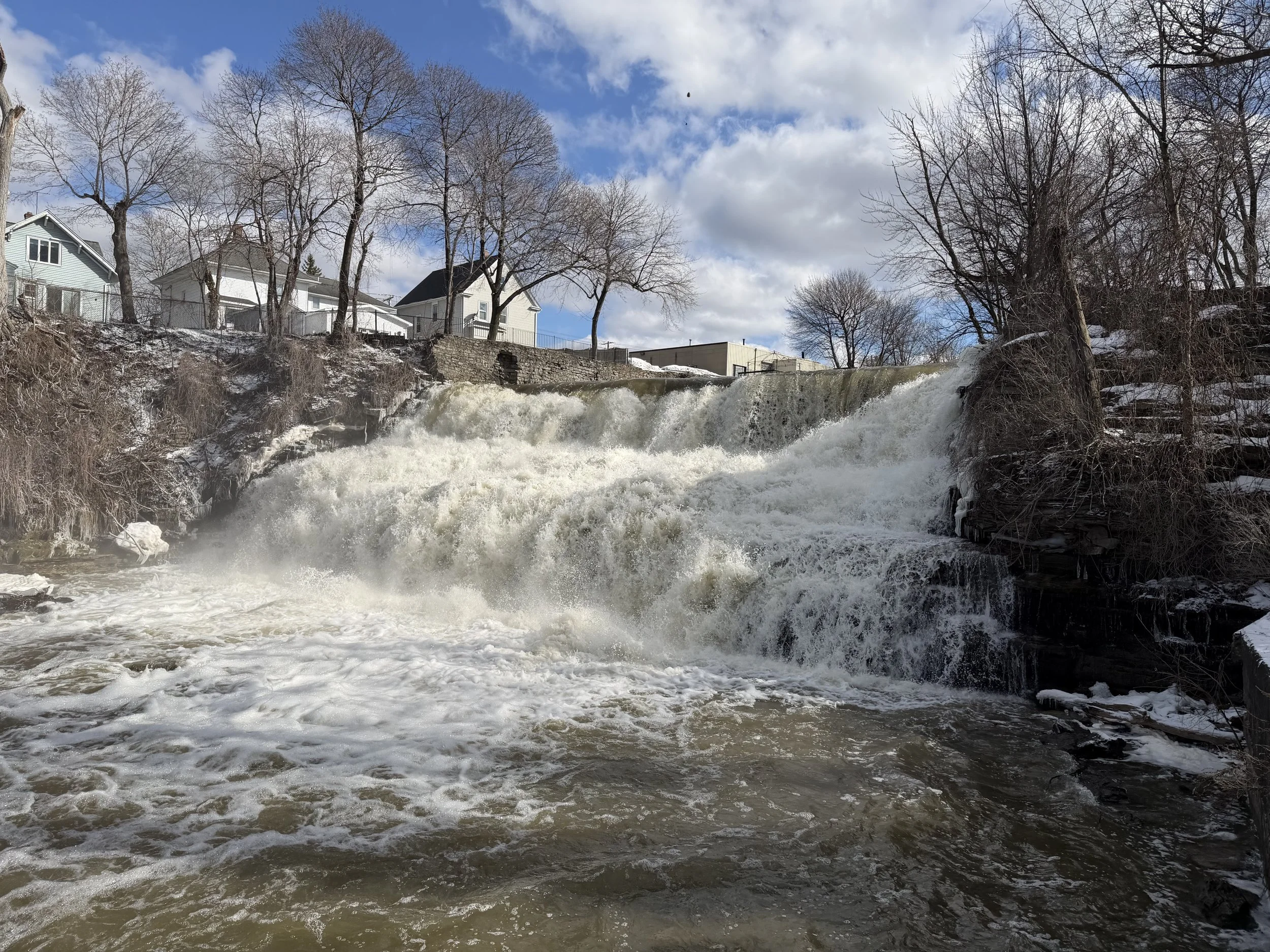 A small waterfall flowing over rocks with houses and trees on a snowy hillside under partly cloudy sky.