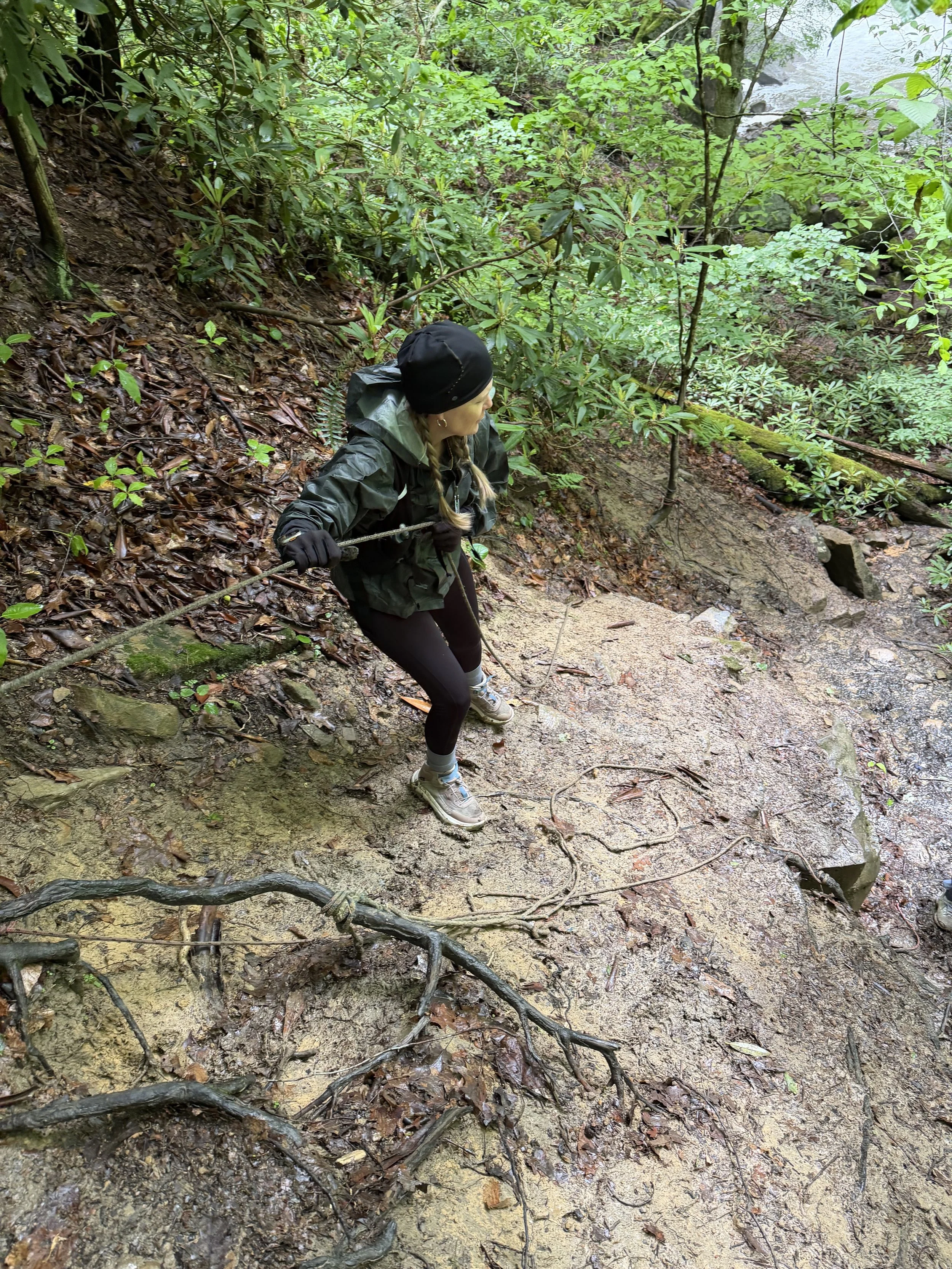 A person in outdoor clothing and gloves climbing a muddy, tree-lined trail with roots and rocks, surrounded by lush green forest.