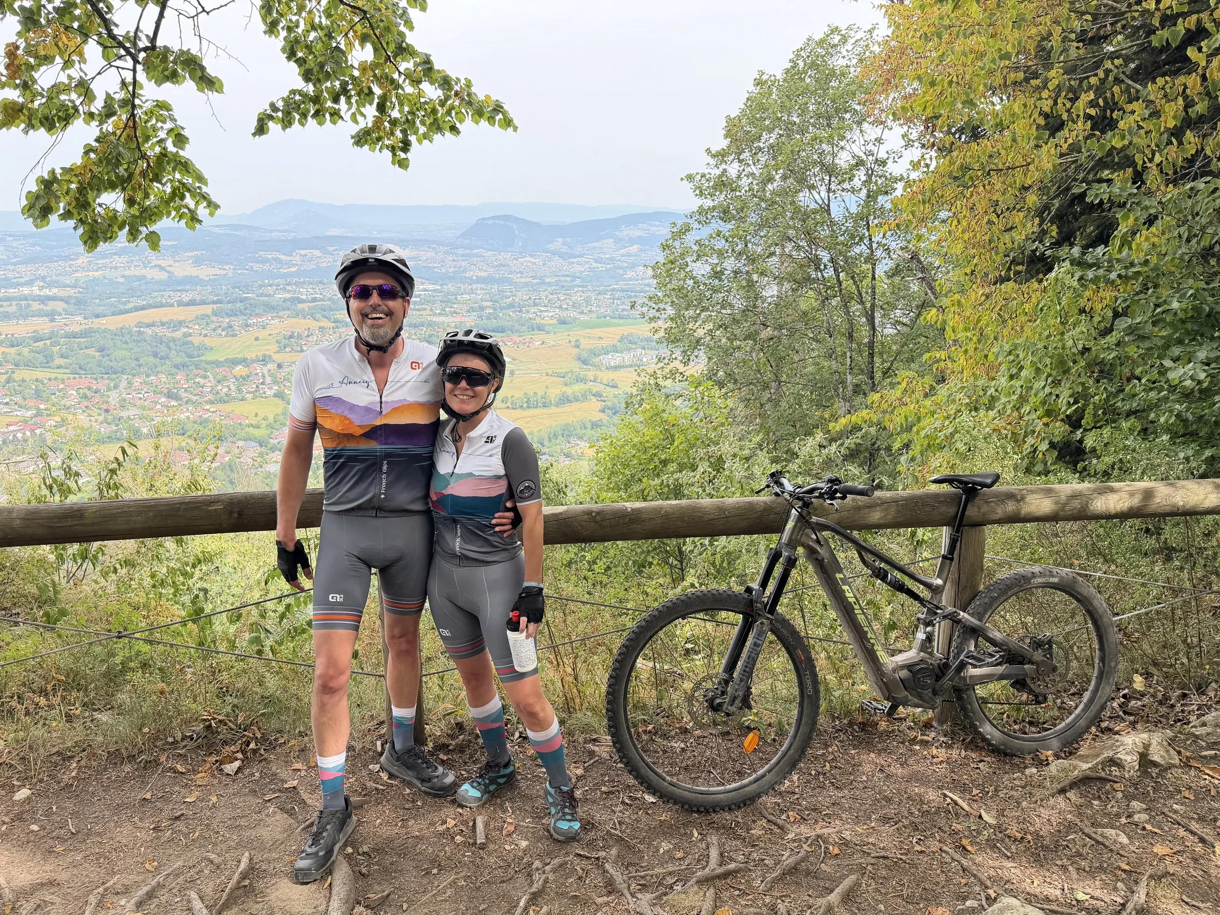 Two happy cyclists in biking gear, wearing helmets and sunglasses, standing together on a trail with a scenic view of a valley and mountains in the background. There is a mountain bike next to them, and they are surrounded by green trees and shrubs.