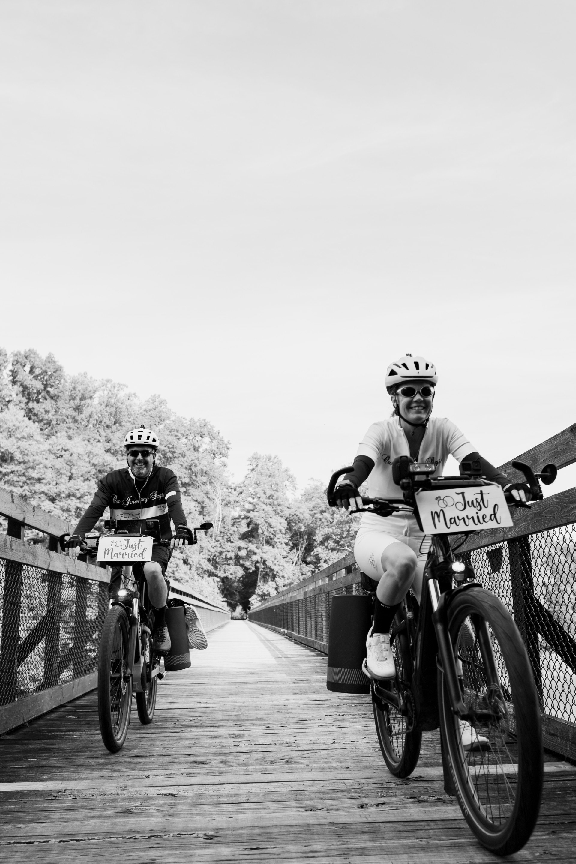 Two women riding bicycles on a wooden bridge, both wearing helmets and sunglasses, each with a sign on their bike that reads 'Just Married.' The women are smiling and appear happy, with trees and a cloudy sky in the background.