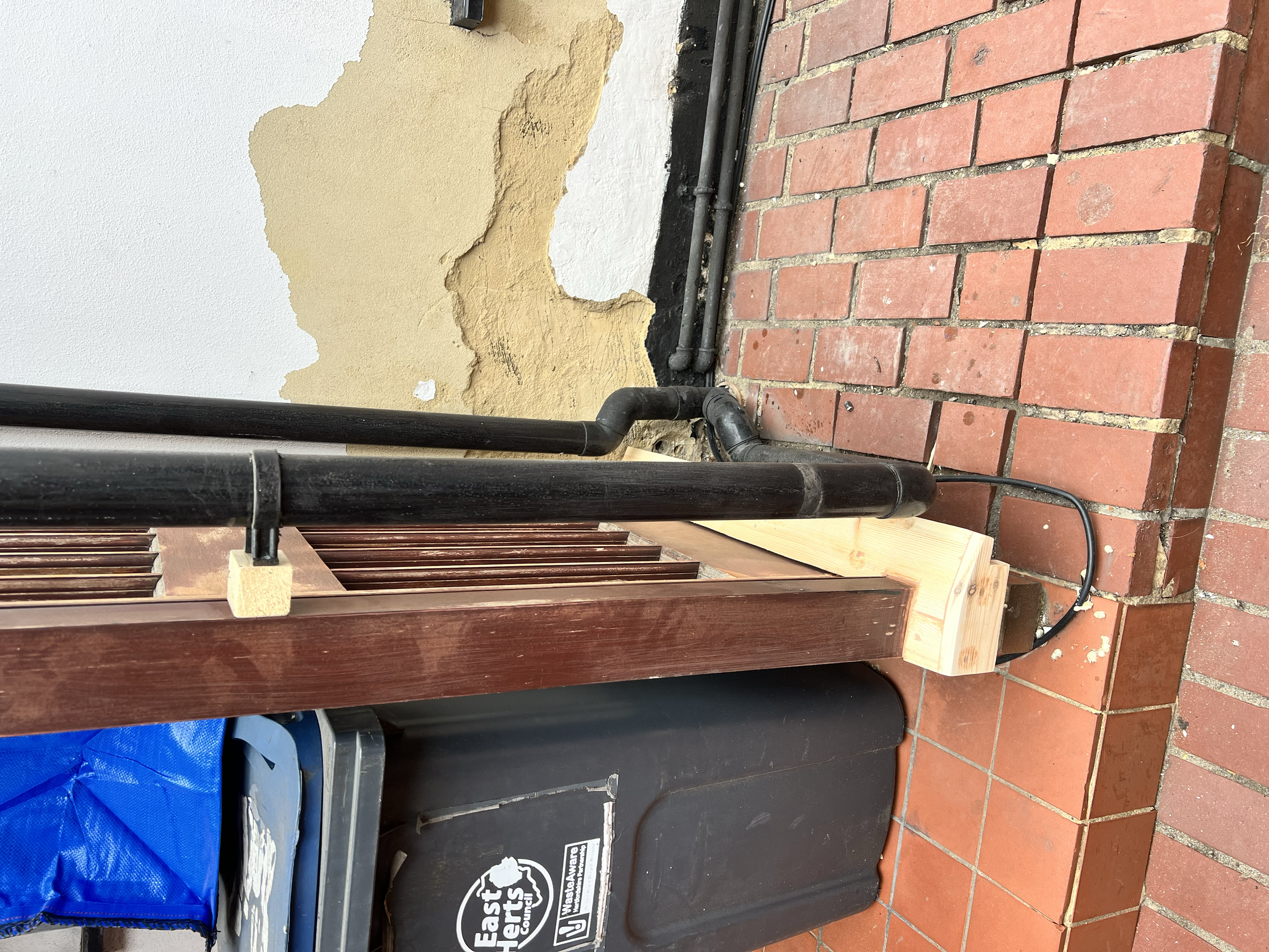 A brick sidewalk next to a wall with peeling paint, and a black trash bin labeled "East Hartford" with a blue bag inside. There are black pipes running along the wall.