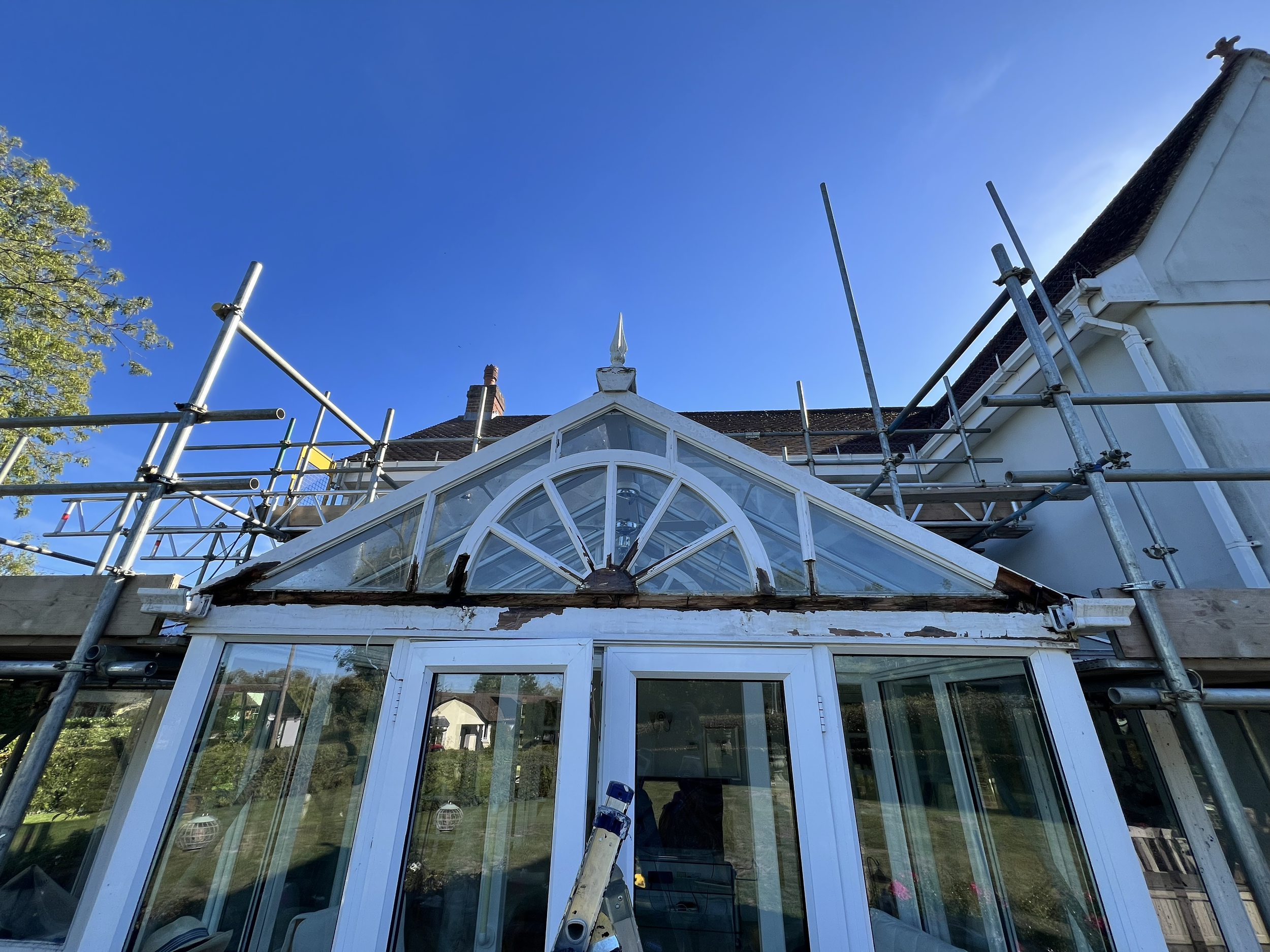Scaffolding around a house with a glass conservatory, a decorative finial on the roof, and a clear blue sky in the background.
