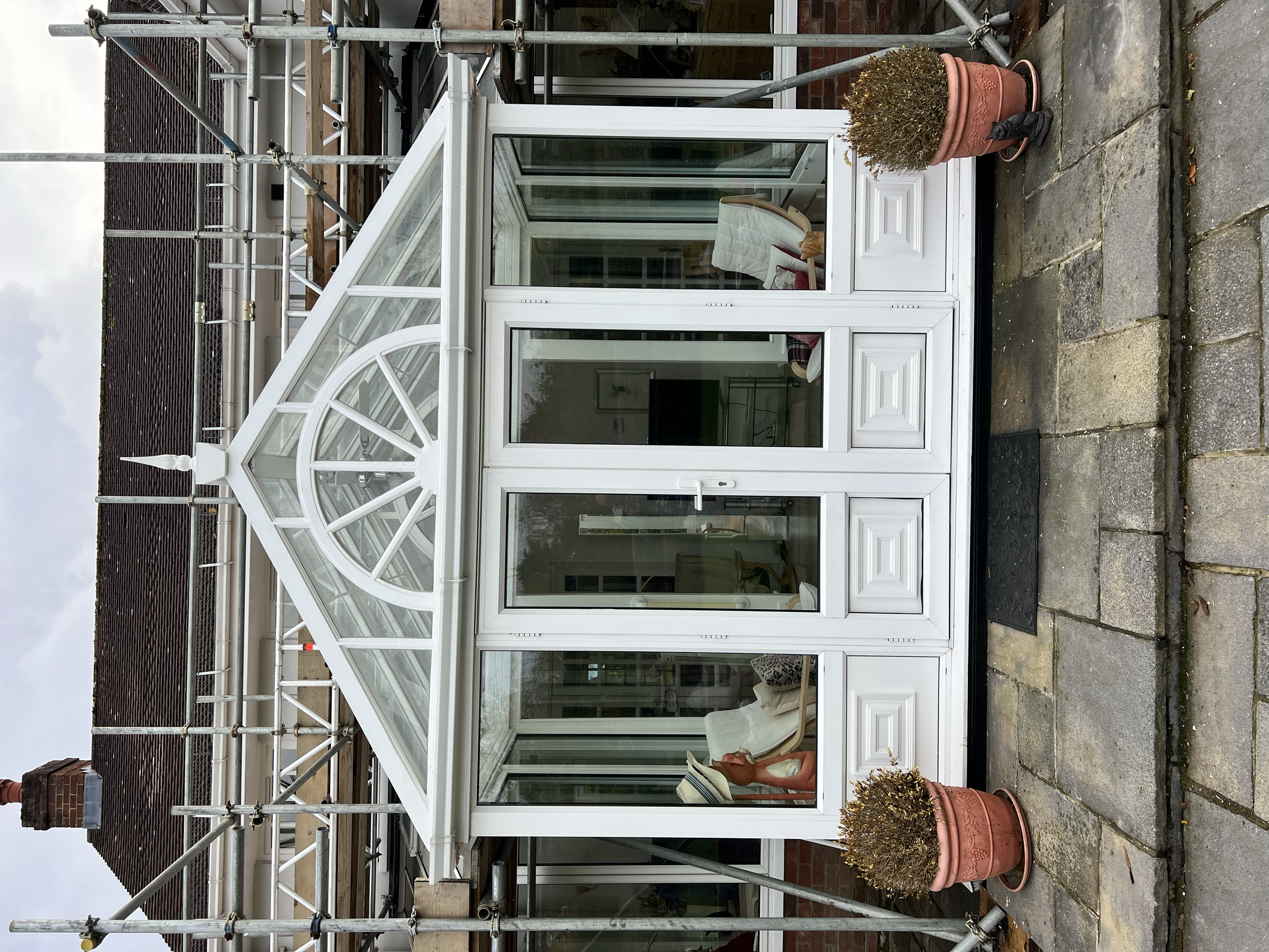 A house under construction with a white conservatory or sunroom. The structure is surrounded by metal scaffolding. Two large potted plants with browned, dried foliage are positioned outside the conservatory on a stone-paved area.