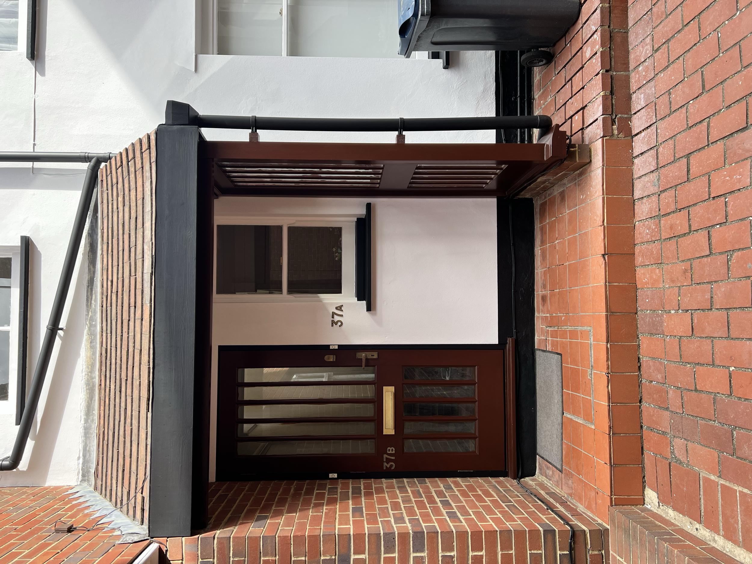 Image of a brick doorway with a small roof overhang, featuring house number 37A and 37B, leading into a building with a glass-paneled door and window.