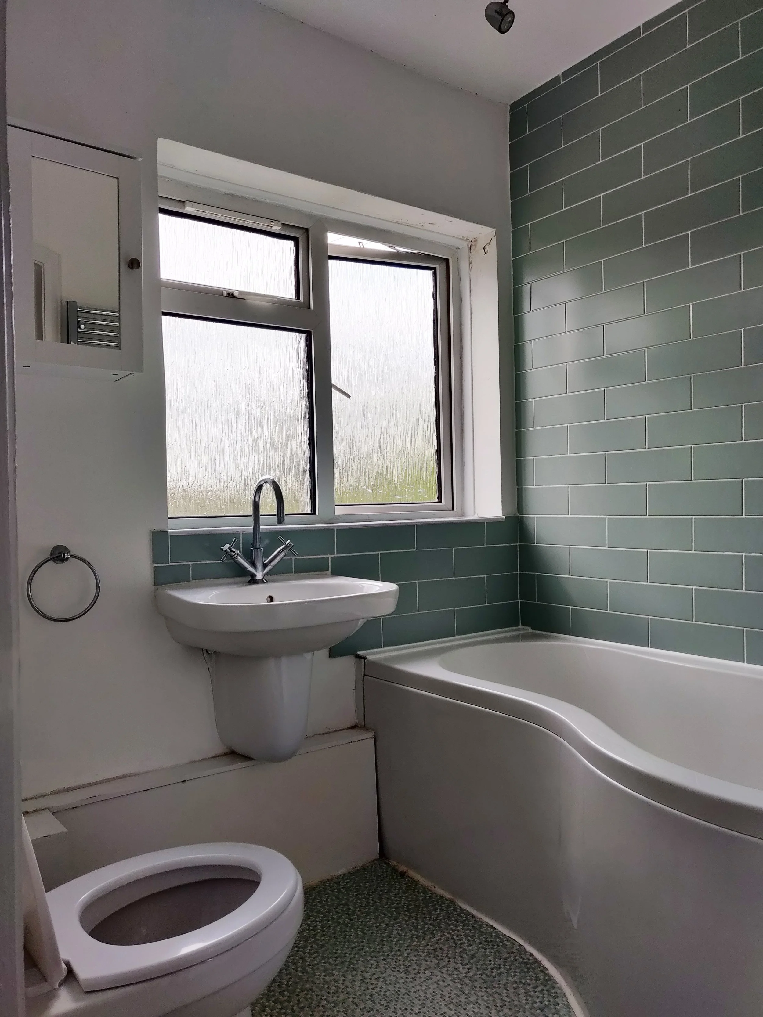 A small bathroom with a window, a white sink, a toilet, a bathtub, green subway tiles on the wall, and a black-rimmed window with frosted glass.