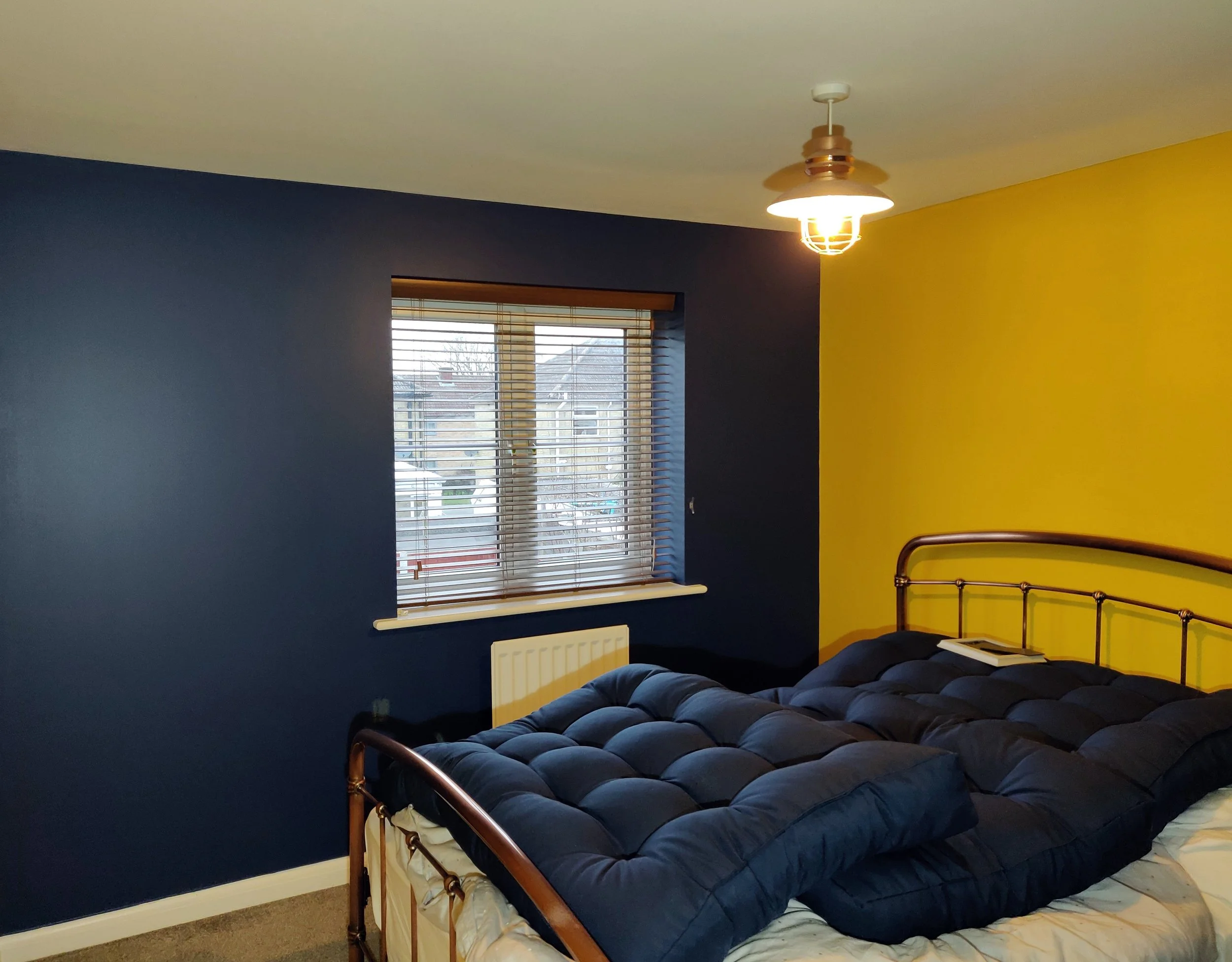 A bedroom with dark blue and yellow walls, a brass bed with a dark blue comforter, a large window with wooden blinds, and a ceiling light fixture.