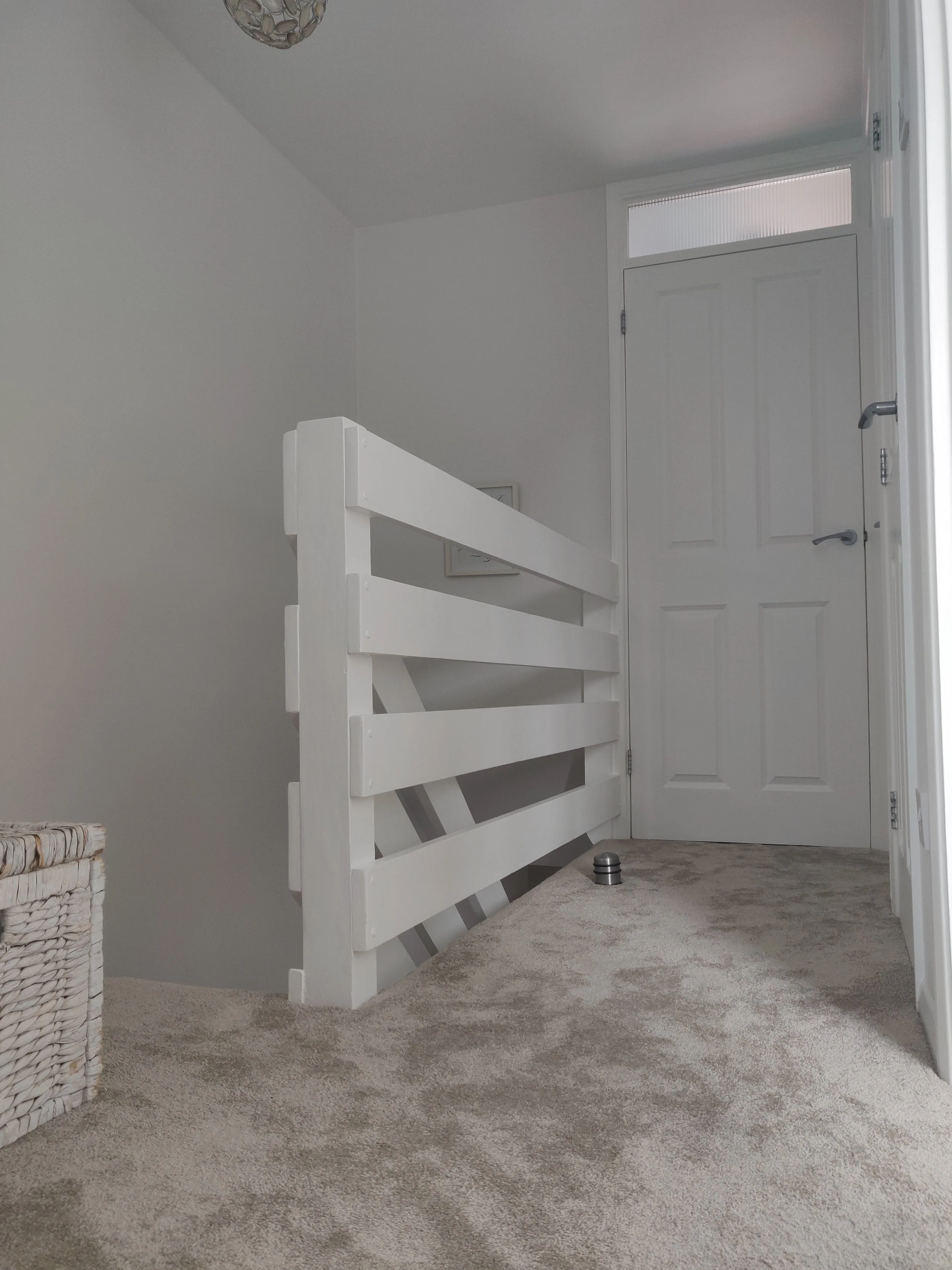 Interior scene with a white wooden bed frame leaning against a white wall, a door with a small window, a woven basket on a beige carpet, and a small metallic object on the floor.