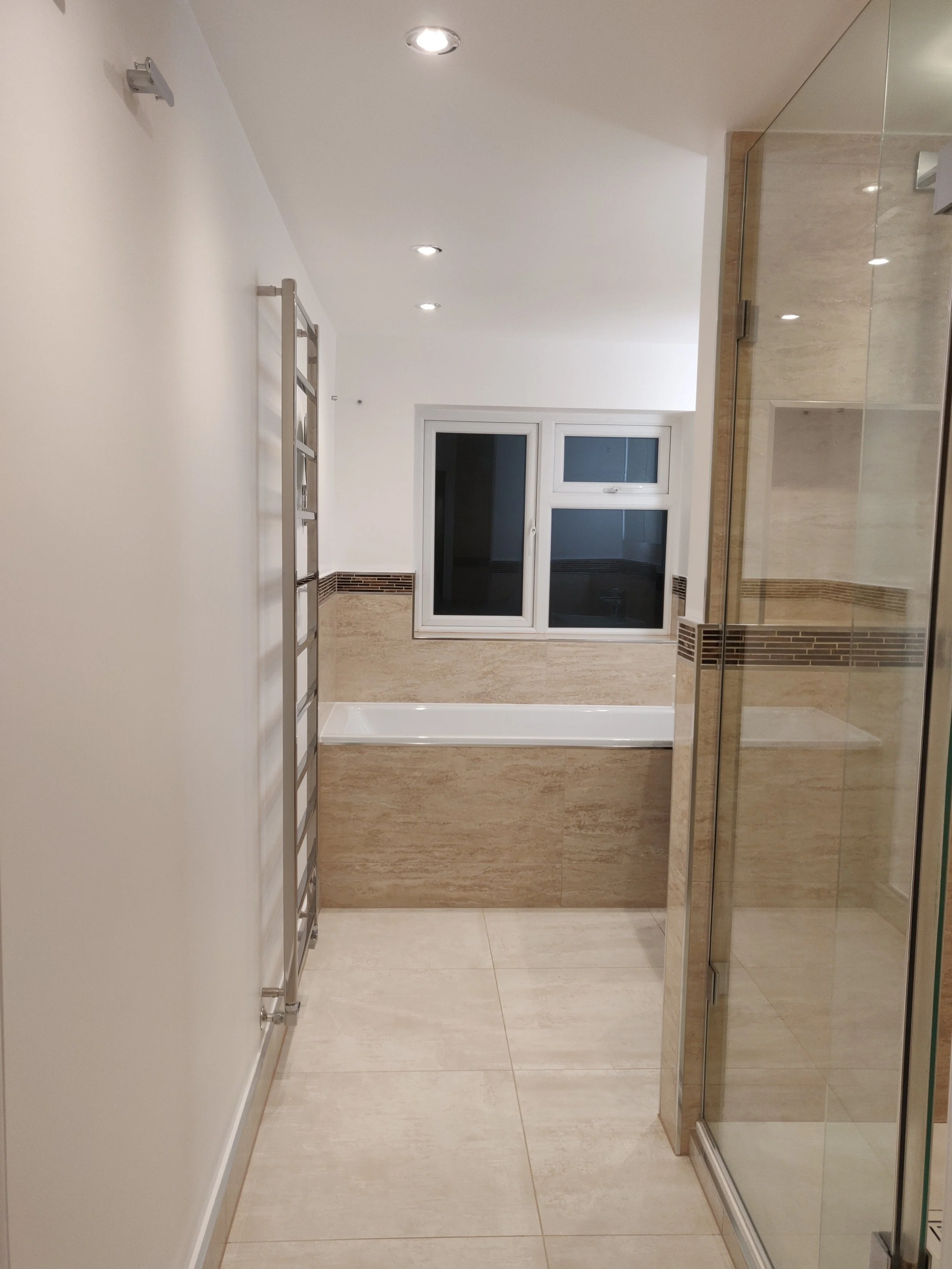 A clean, modern bathroom with beige tiled floors and walls, a white window, and a glass shower enclosure on the right. There is a silver towel rack mounted on the left wall and recessed ceiling lights.