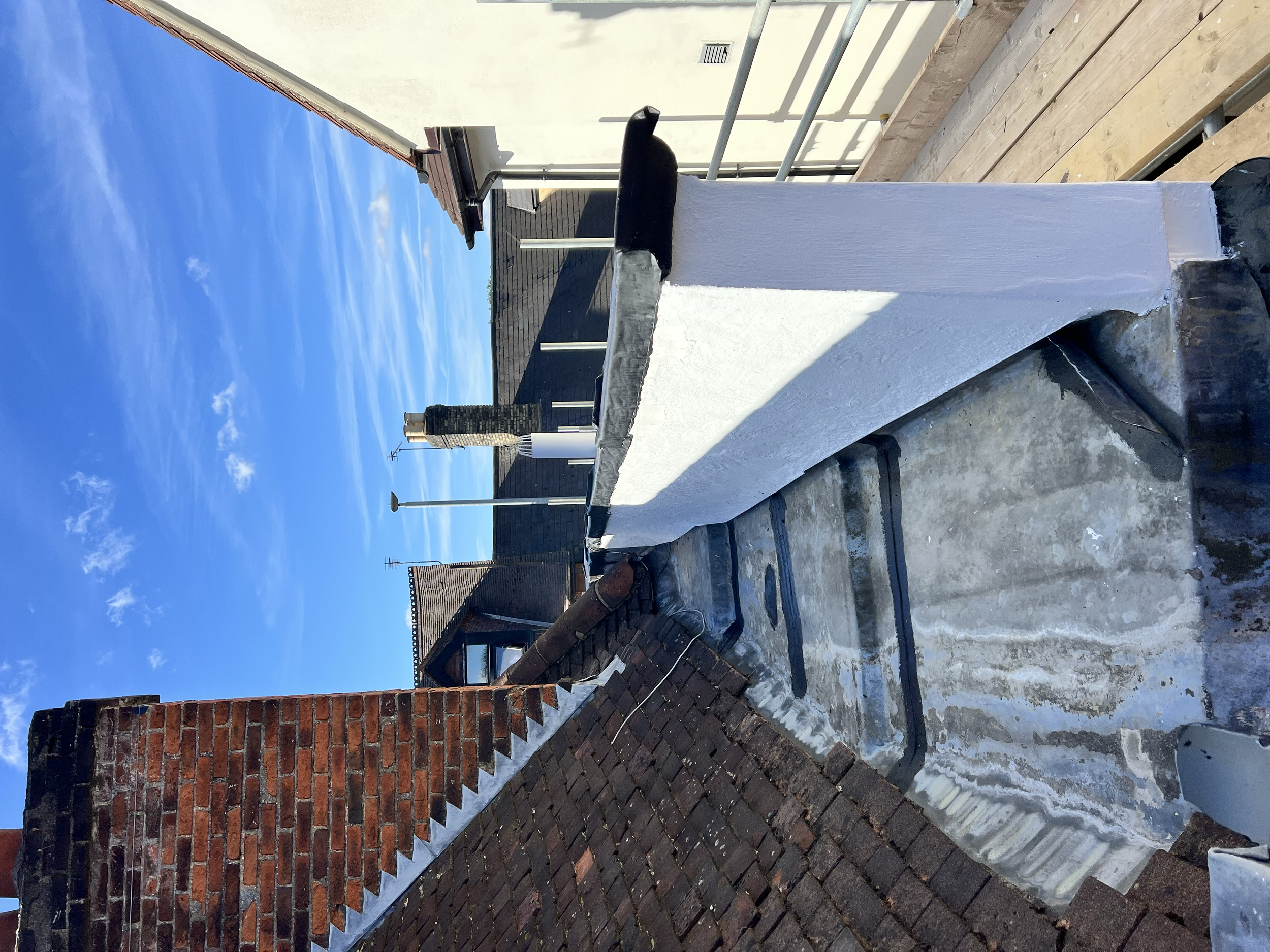View of rooftops and chimneys against a bright blue sky with some wispy clouds, showing part of a building's rooftop with a chimney, brickwork, and a white chimney pipe.