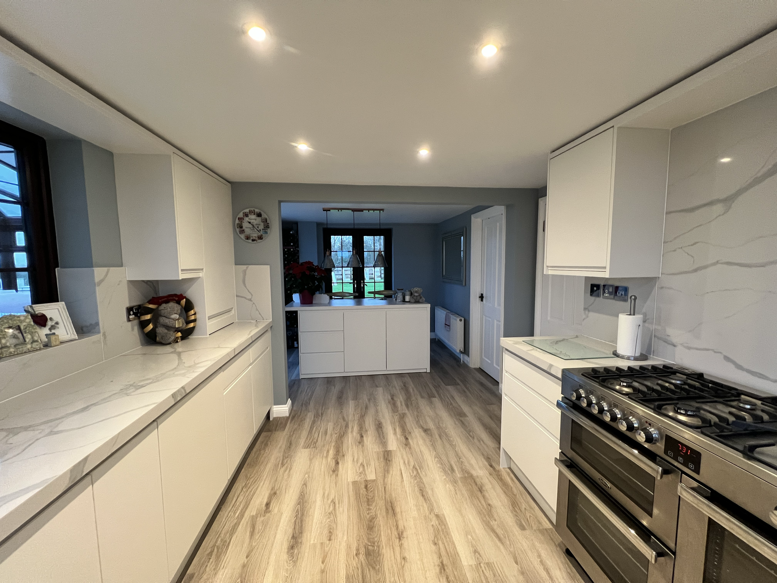 A modern kitchen with white cabinets, marble countertops, a gas stove, wooden flooring, and recessed lighting, with a view of a dining area with hanging pendant lights and large windows in the background.