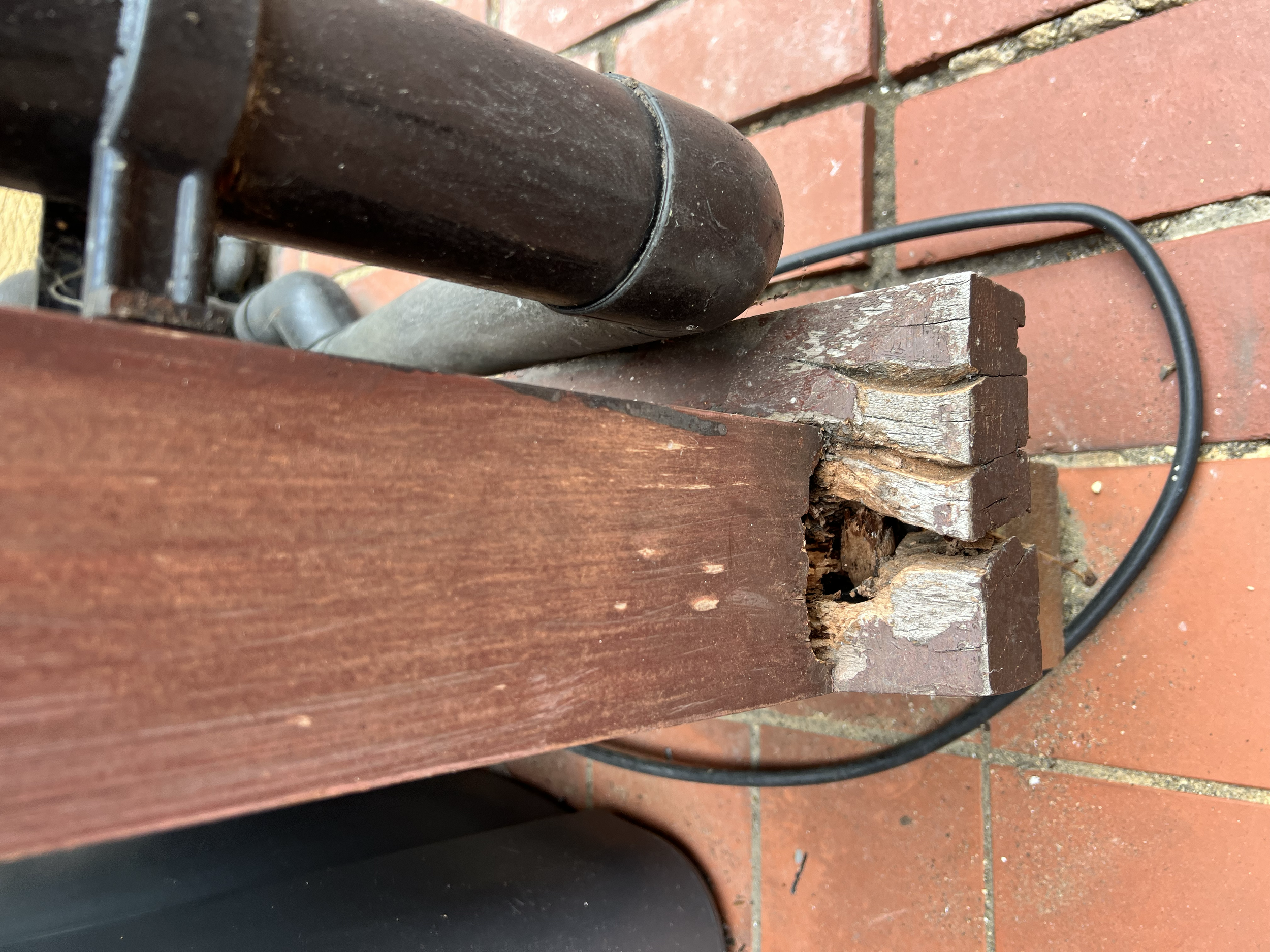 Close-up of a part of a wooden bench, showing the armrest and a section of the seat with red paint peeling off, on a brick floor.