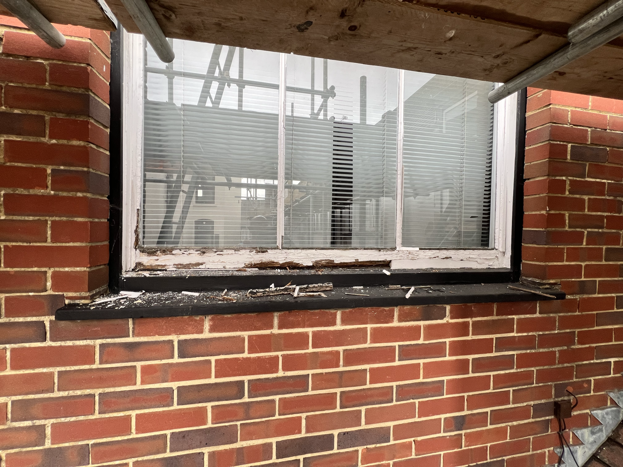 A window with a white frame and horizontal blinds, set in a red brick wall. The window sill has peeling paint and some debris. The reflection in the window shows scaffolding outside.