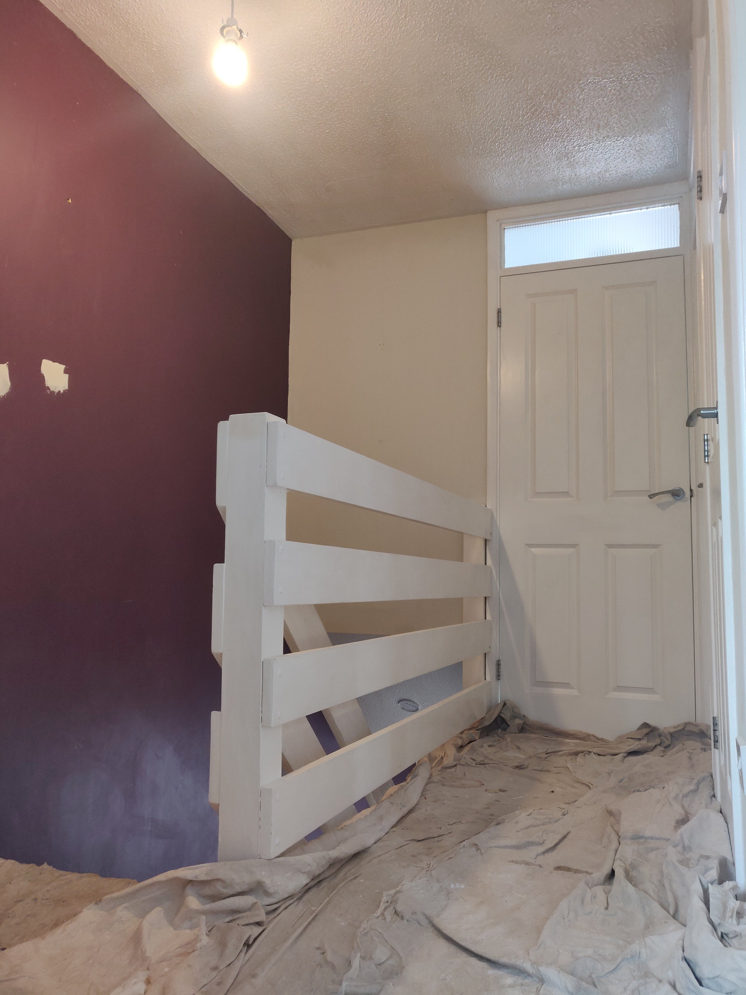 Interior of a room under renovation with a white wooden staircase, a closed white door, partially painted walls with one maroon wall and the rest in light cream, a ceiling light bulb, and painter's drop cloth covering the floor.