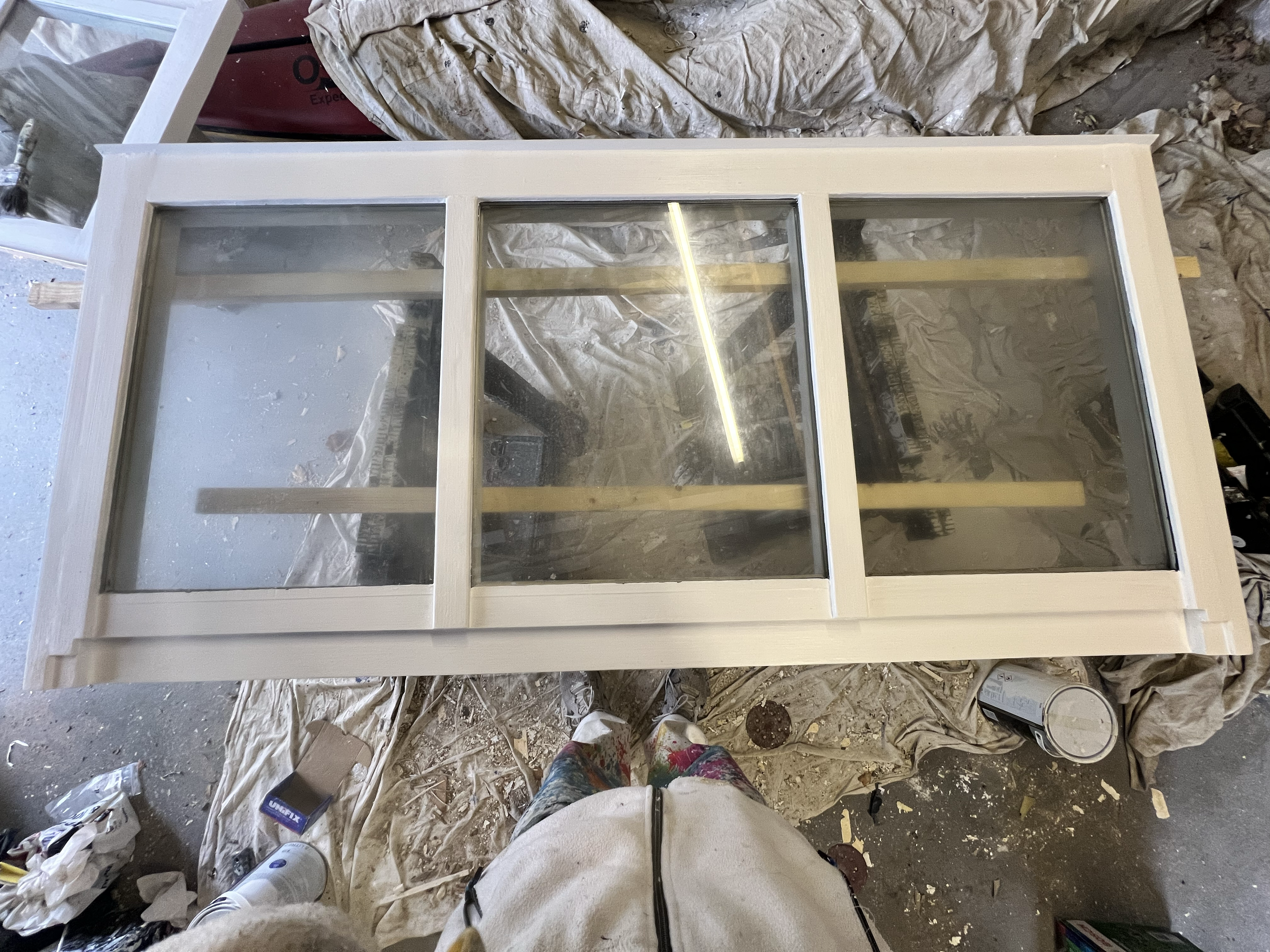 This image shows a partially assembled white wooden frame with large glass panels, possibly a window or door, set in a workshop or construction area. The surrounding area is cluttered with plastic sheeting, paint cans, tools, and other construction m