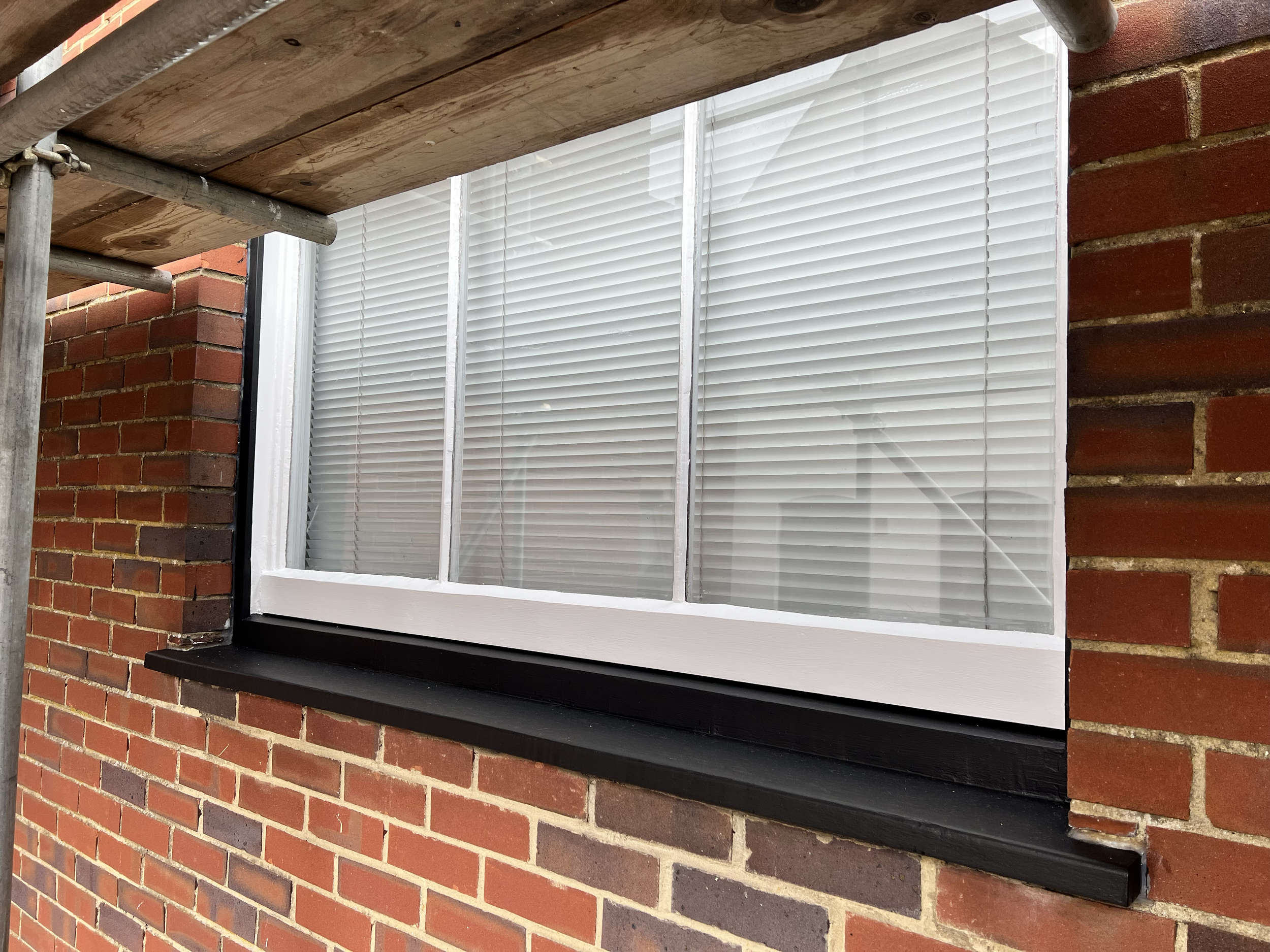 Close-up of a window with white blinds on a brick building, framed by a black window sill and with part of a wooden scaffolding in the upper left corner.