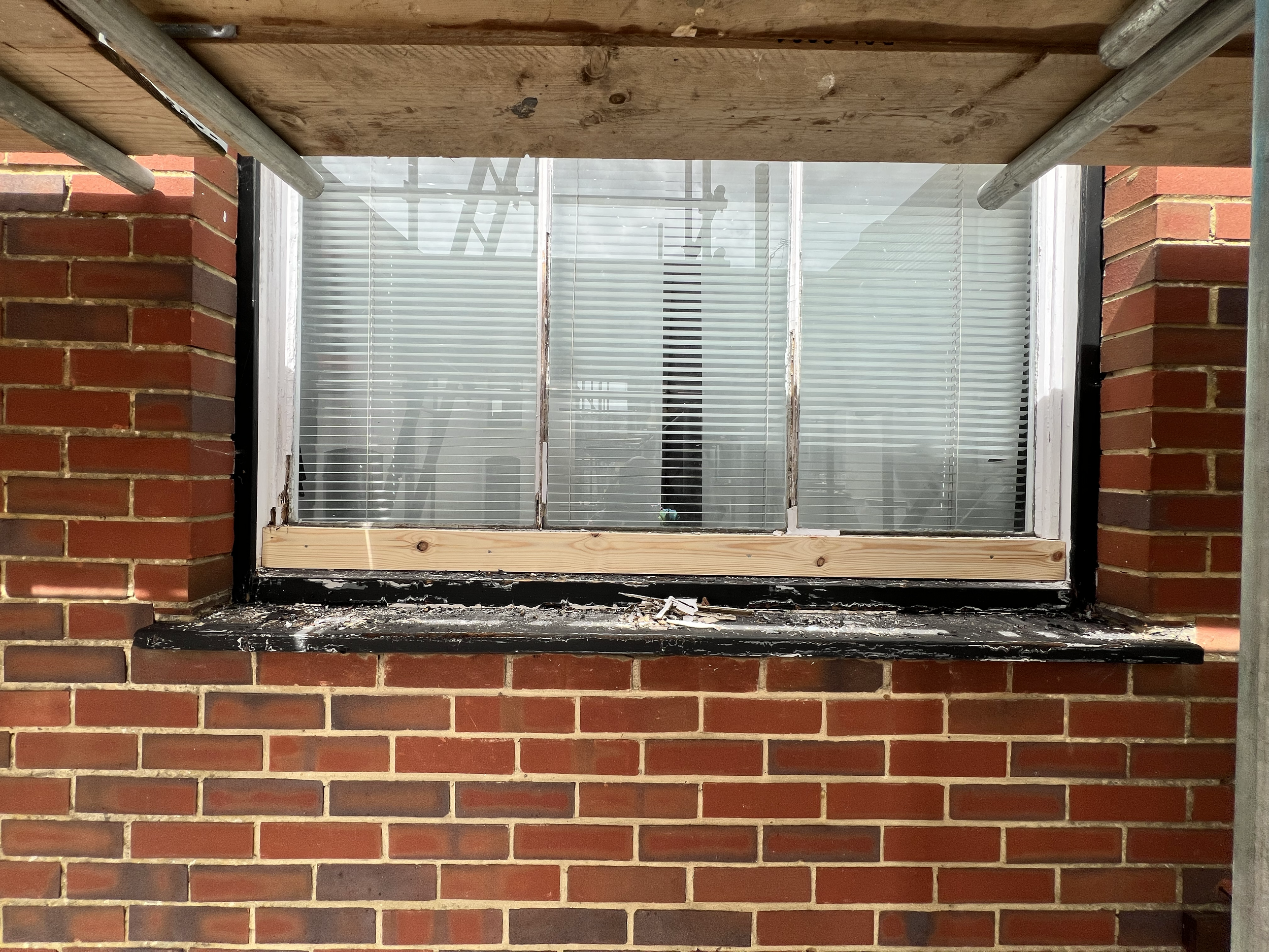 A row of red bricks with a window frame under construction, surrounded by metal scaffolding, and a wooden board above the window.