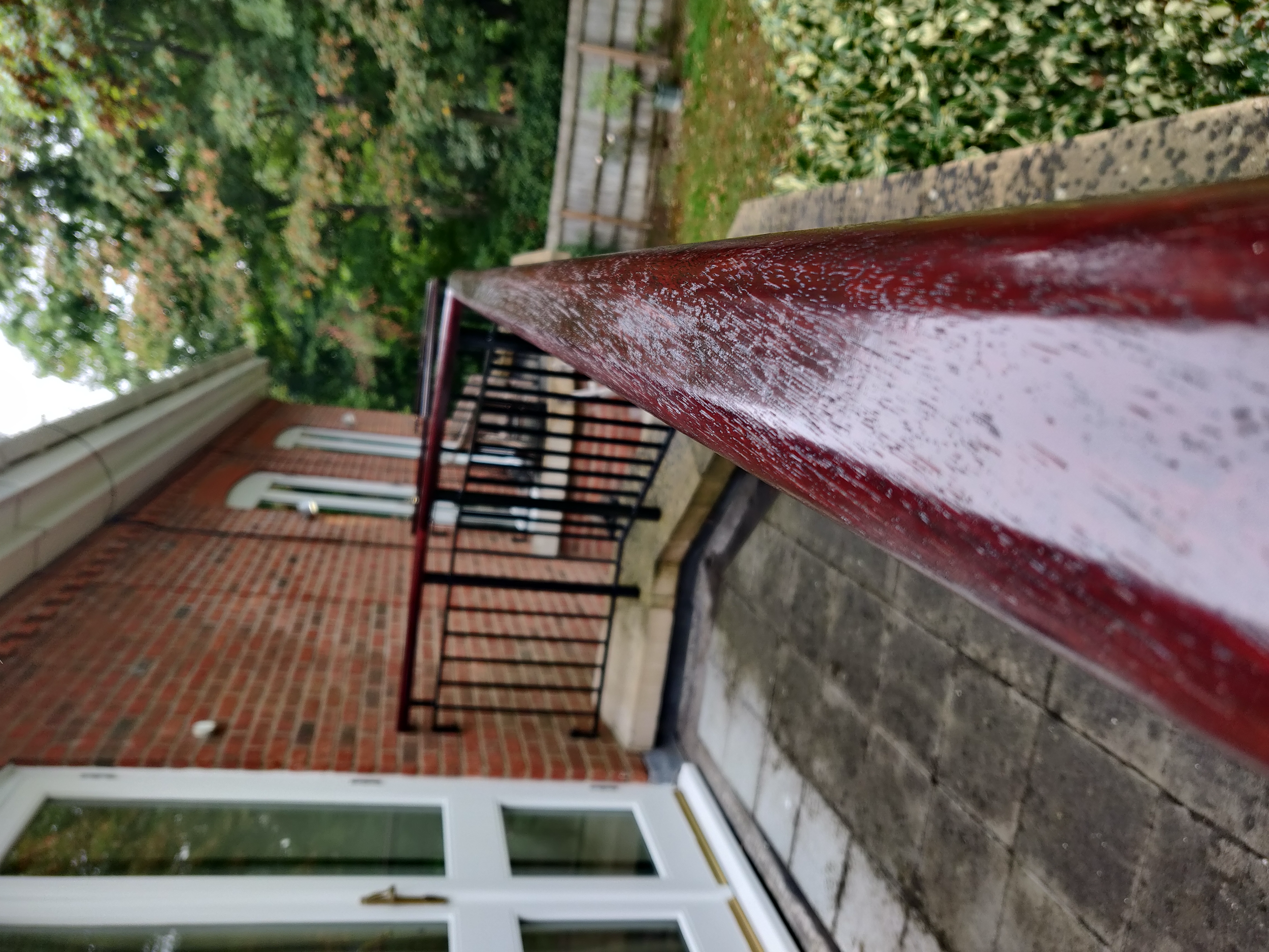 A close-up of a red railing on a balcony, with a view of trees and a brick building with white-framed windows in the background.