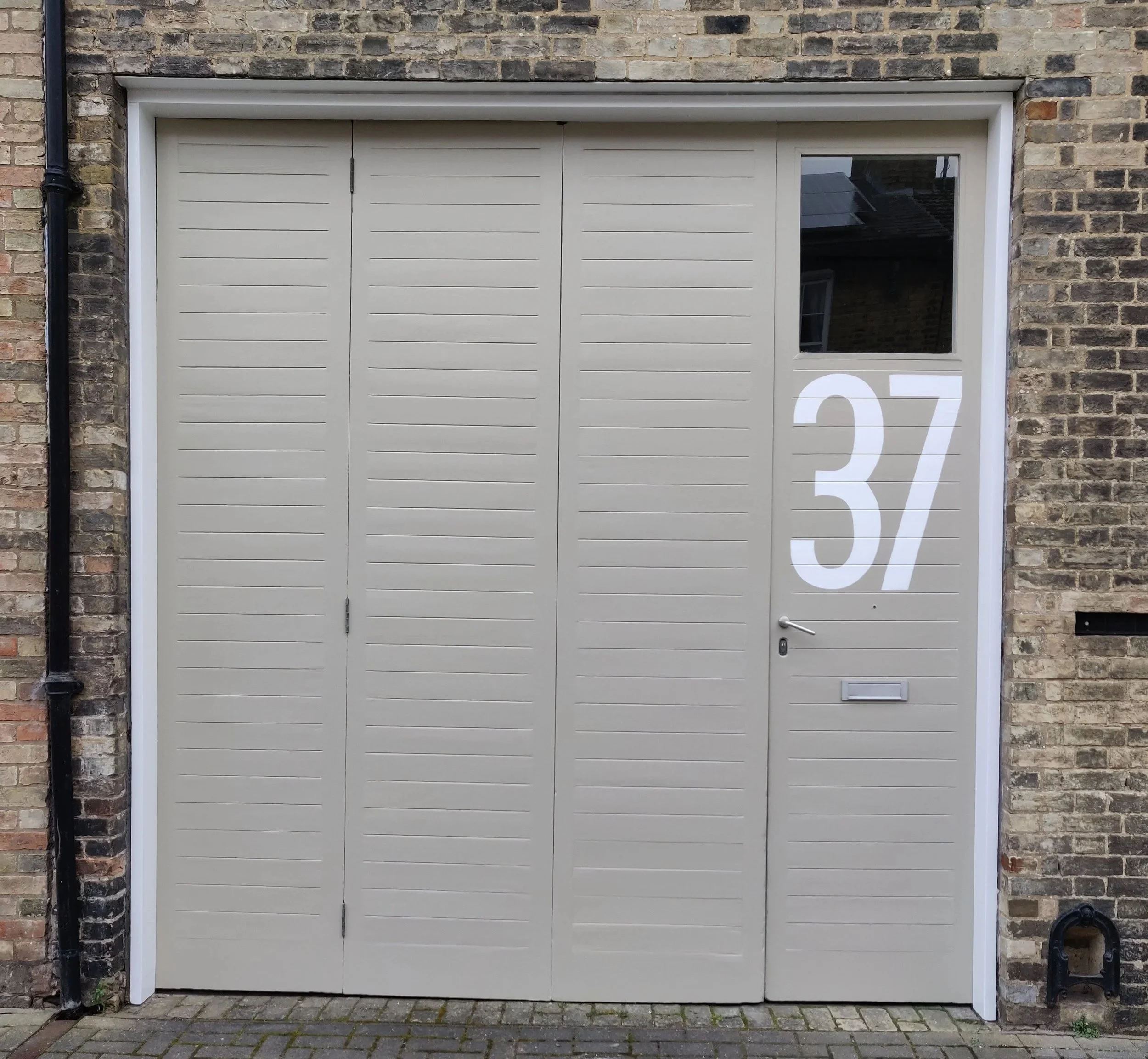 A beige-colored garage door with horizontal lines, set in a brick wall with a black gutter pipe on the left side, and a single glass window to the right of the door showing part of a building interior. White trim surrounds the door and window, and the number 37 is partially visible at the bottom right corner.