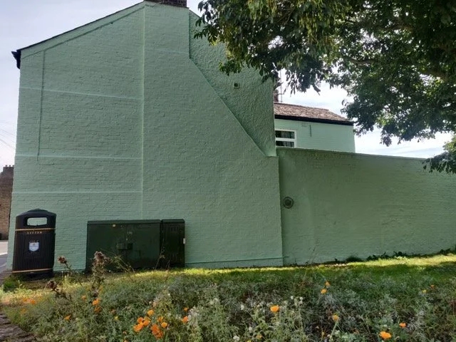 Side view of a mint green painted building with a section slanting downward, a tree casting shade, a trash can, and some orange flowers near the grass in the foreground.