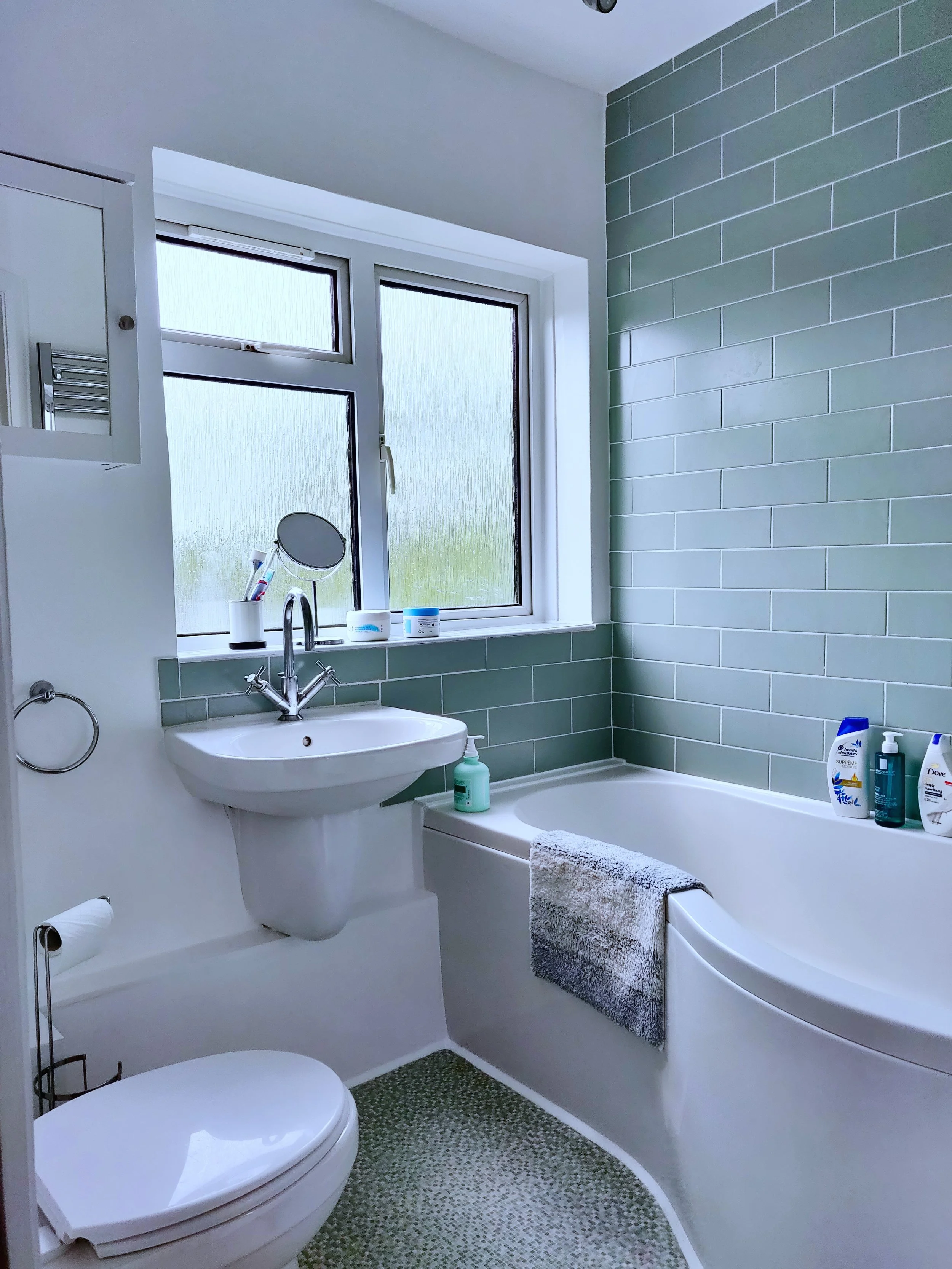 A modern bathroom with a window, a white toilet, a small white sink, a mirror, and toiletries on the bathtub ledge. The wall features green tiles, and there is a towel hanging on the side of the bathtub.