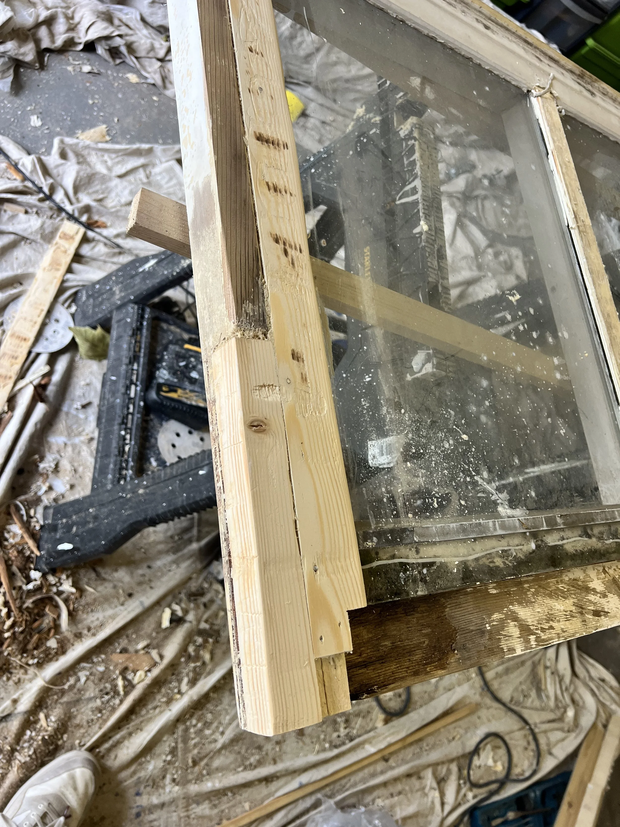 A partially constructed wooden frame with a glass panel, surrounded by construction tools, debris, and work materials in a workshop or construction site.