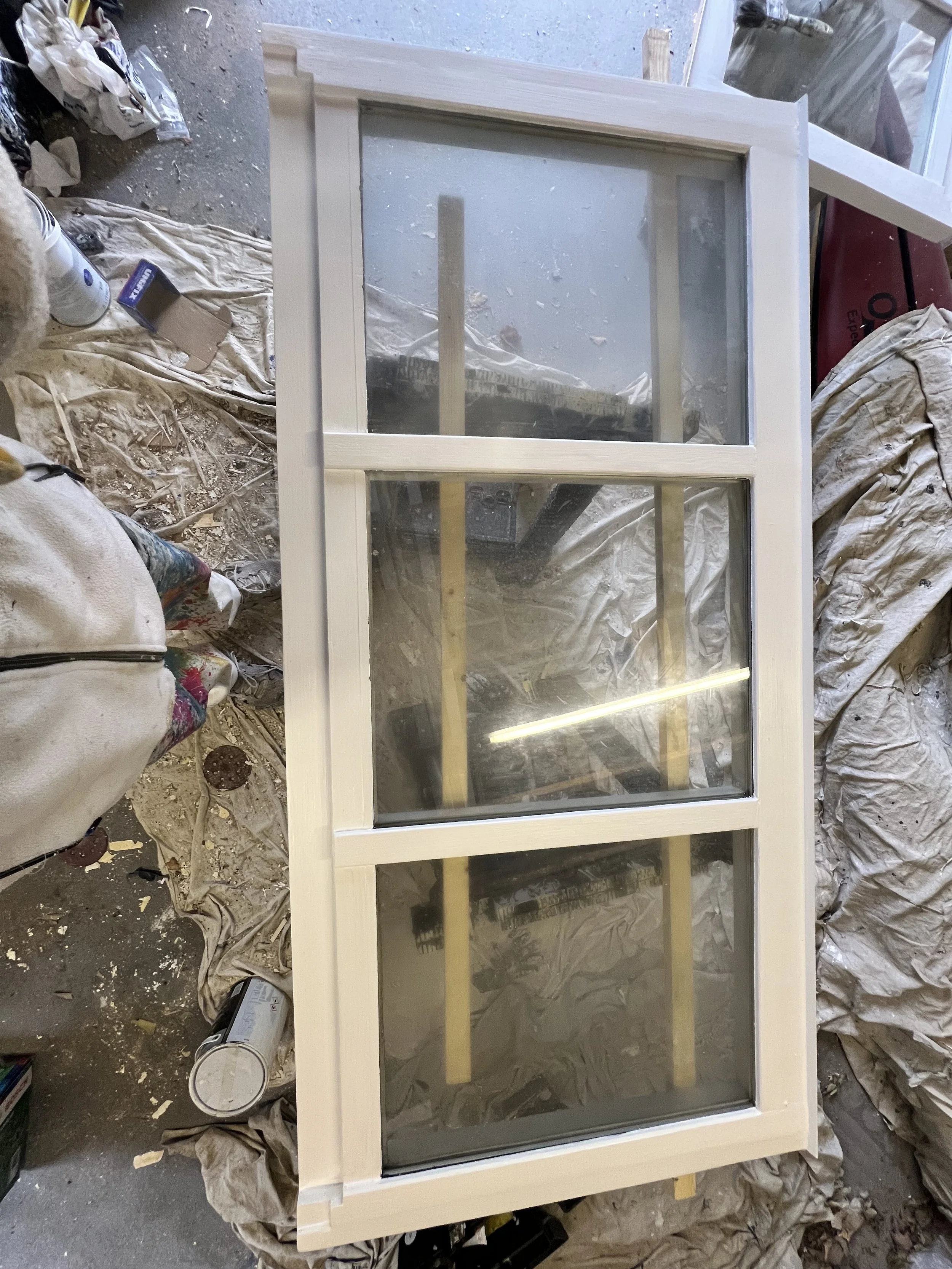 A white wooden window frame with four glass panes, leaning against a wall covered in construction supplies, plastic, and debris.