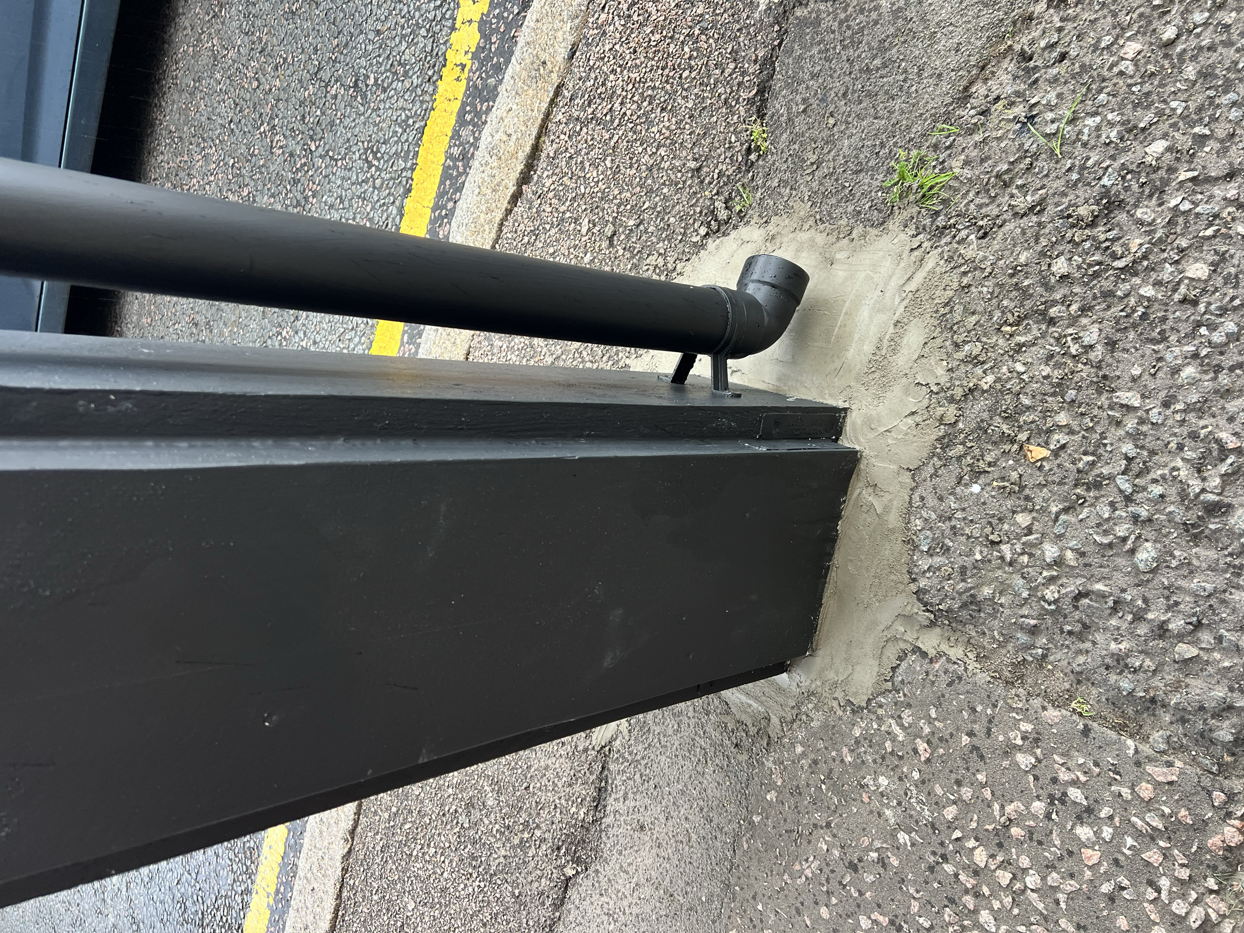 Close-up view of the bottom of a bus stop made of black metal, with a curved black pipe near the sidewalk. The sidewalk is a mix of concrete and gravel, with small patches of grass growing through cracks. There is a yellow line painted on the asphalt