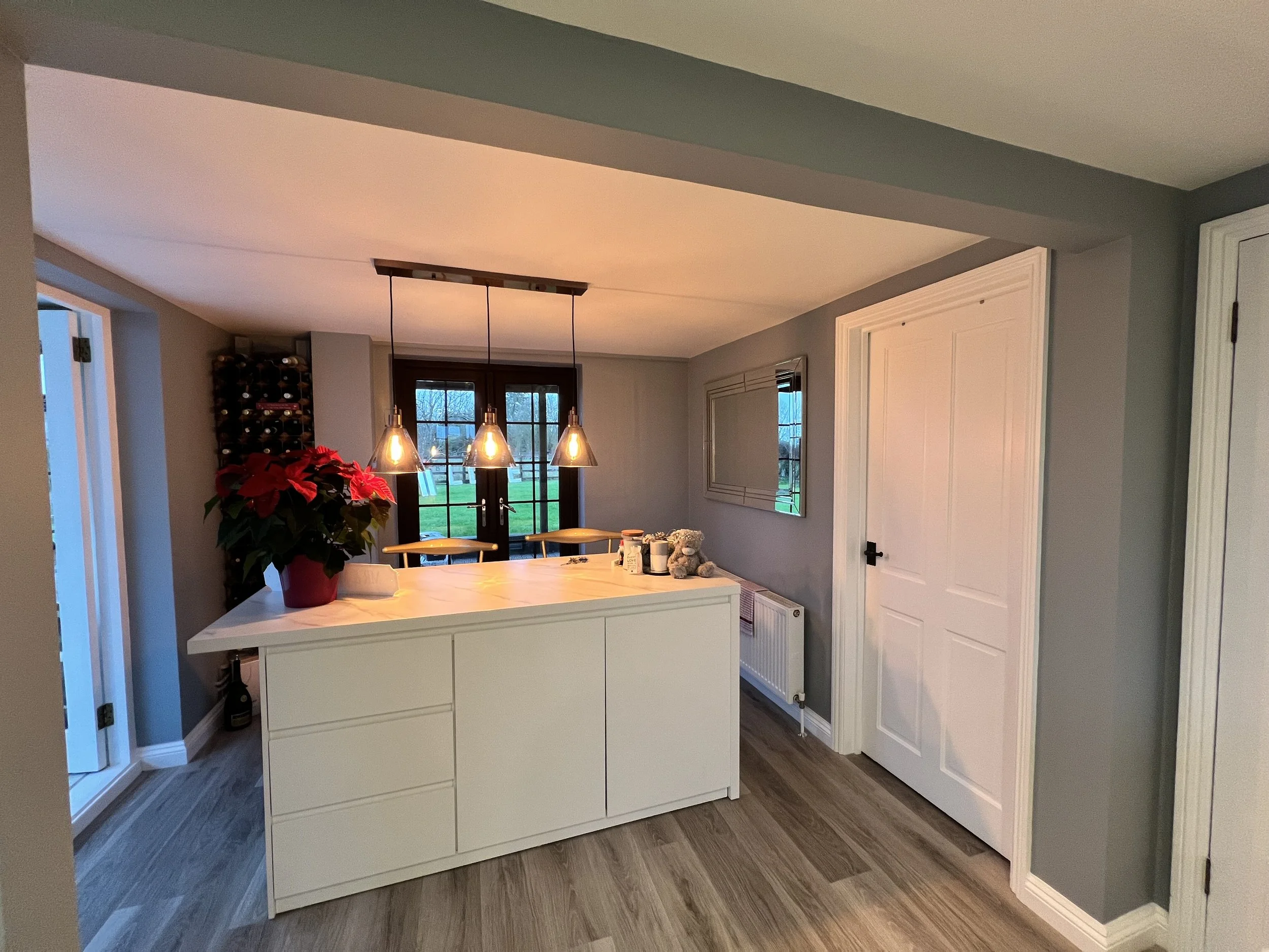 A dining room in a house with a white kitchen island with drawers, a poinsettia plant in a red pot, a window with a view of a green yard, and a cluster of hanging pendant lights.