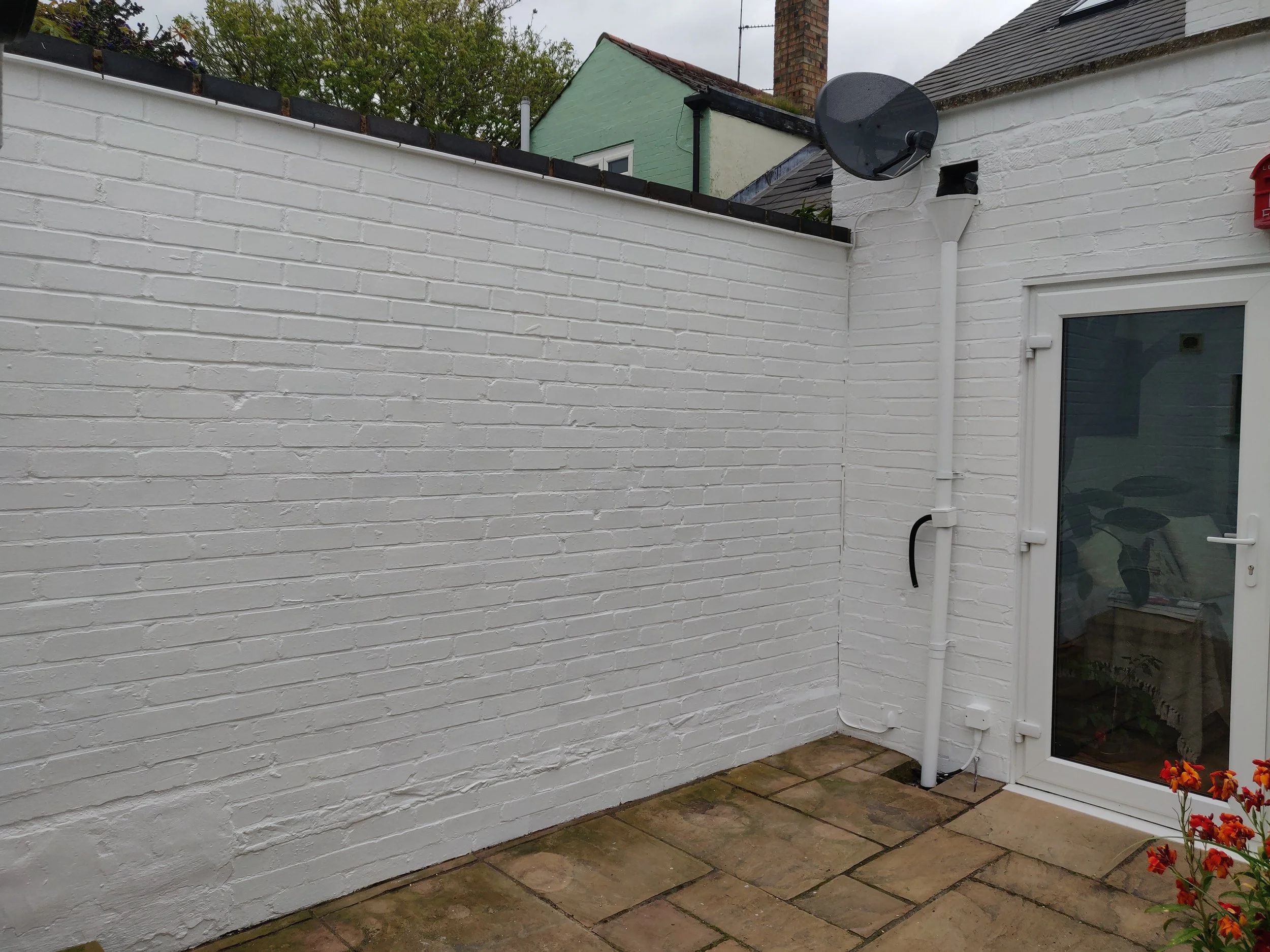 Backyard patio with white painted brick wall, tiled paving, and a glass door. There is a satellite dish and a chimney visible on neighboring roofs. Some flowers are seen in the bottom right corner.